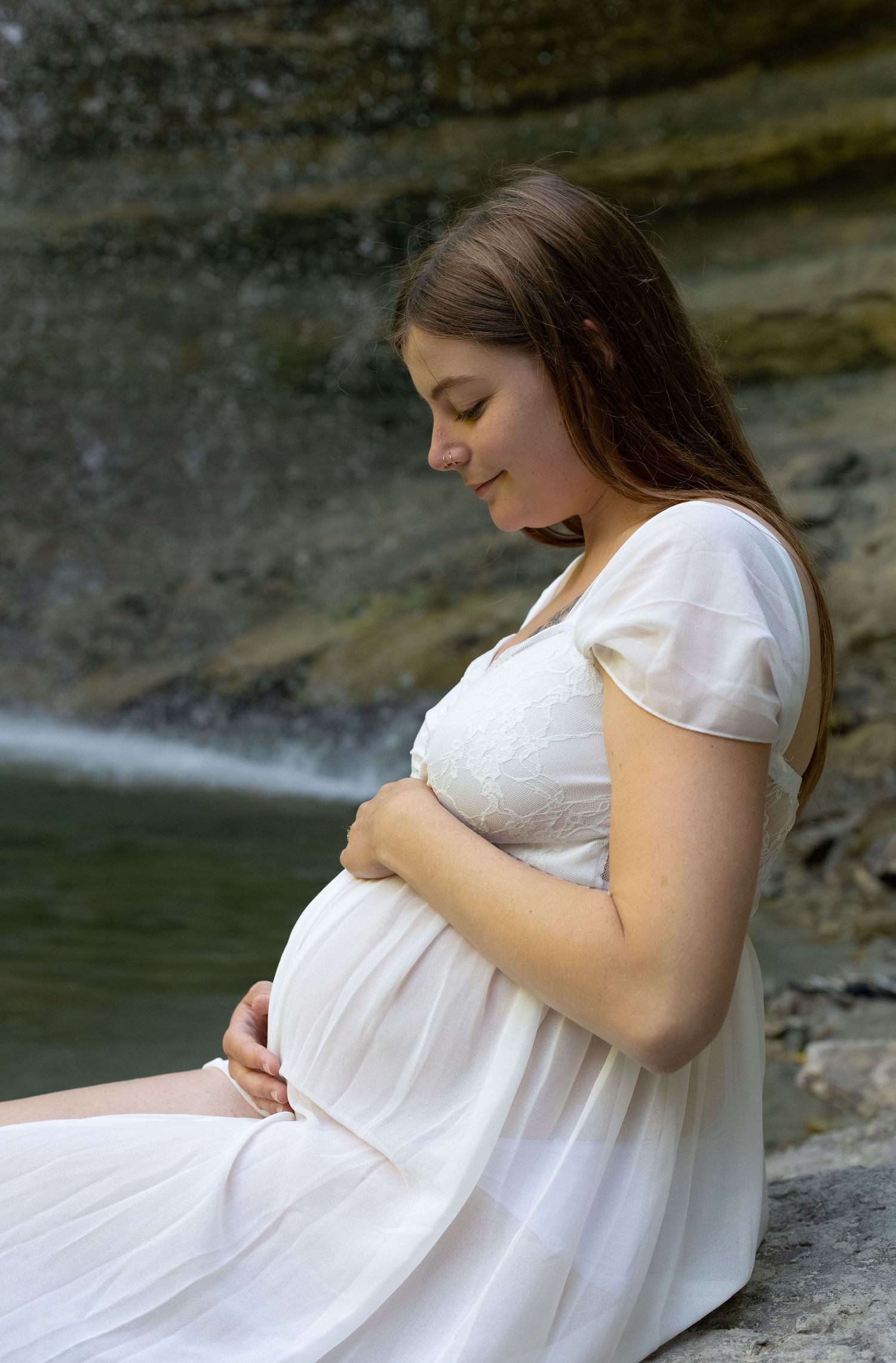 Pregnant woman in white dress sitting near waterfall, smiling and cradling her baby bump.