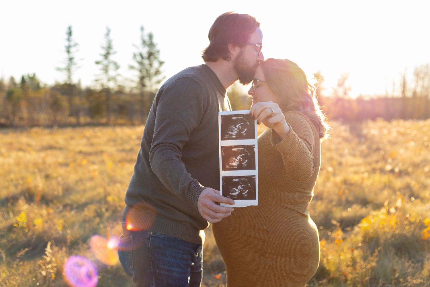 Couple kissing at sunset in a field, holding ultrasound photos, announcing their pregnancy outdoors.