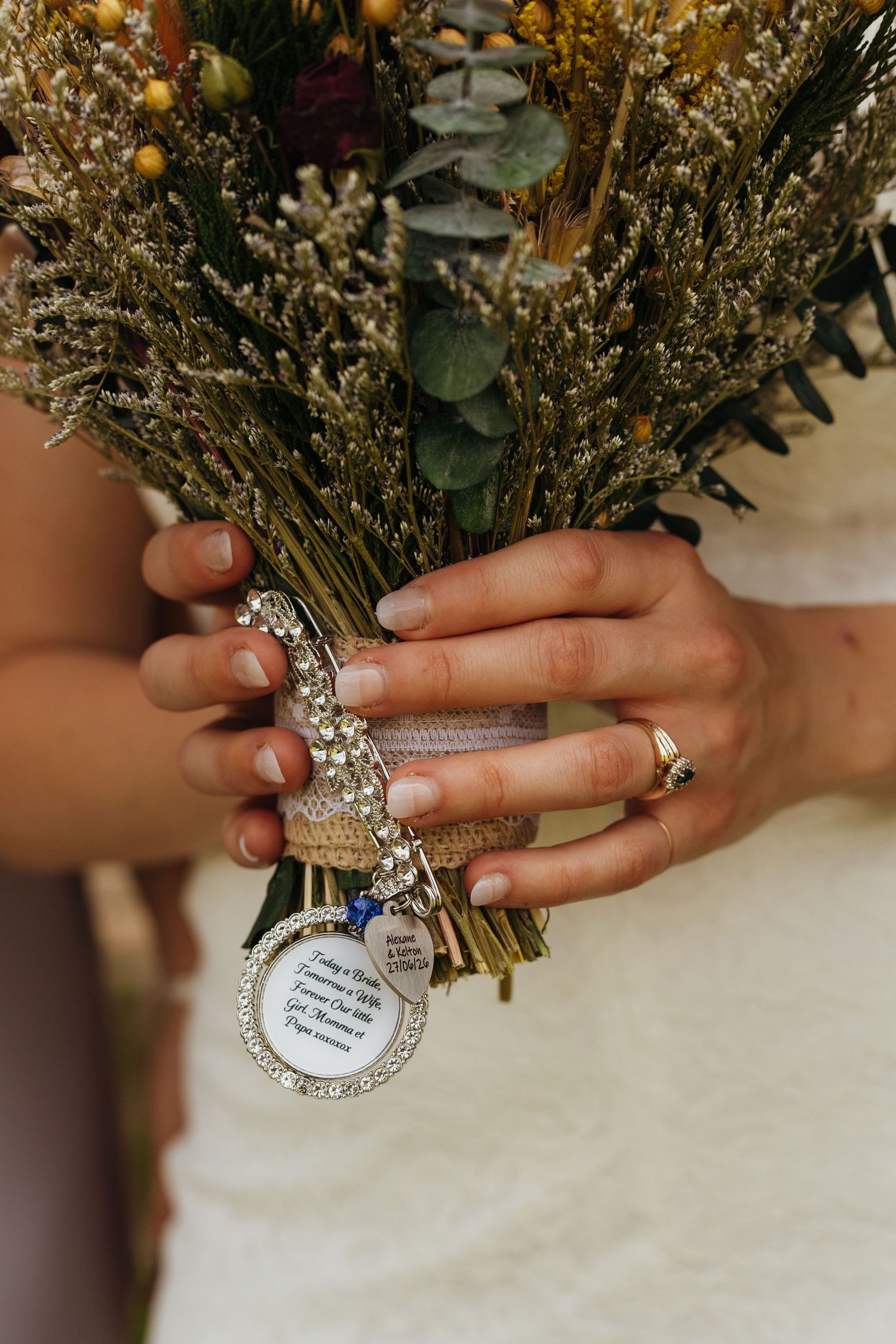 Bride holding a wildflower bouquet with a decorative memorial charm and rhinestone brooch, wearing a diamond ring.