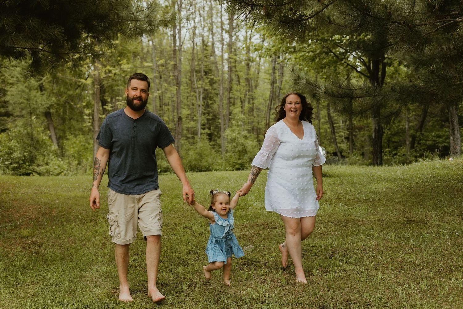 Family of three walking barefoot on green grass with tall trees in background, parents holding toddler girl's hands.