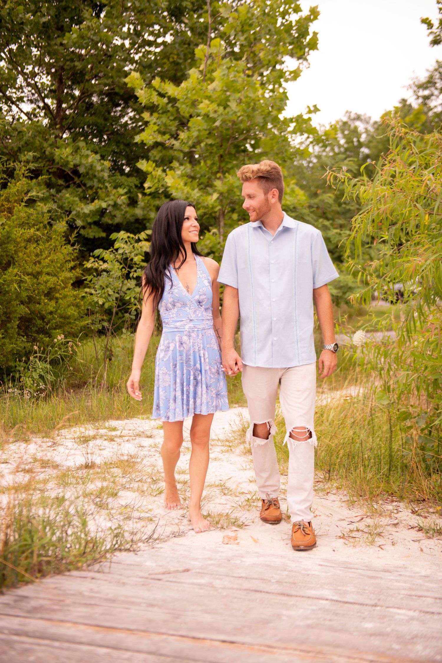 Couple walking hand in hand on sandy path surrounded by lush greenery, woman barefoot in blue dress.