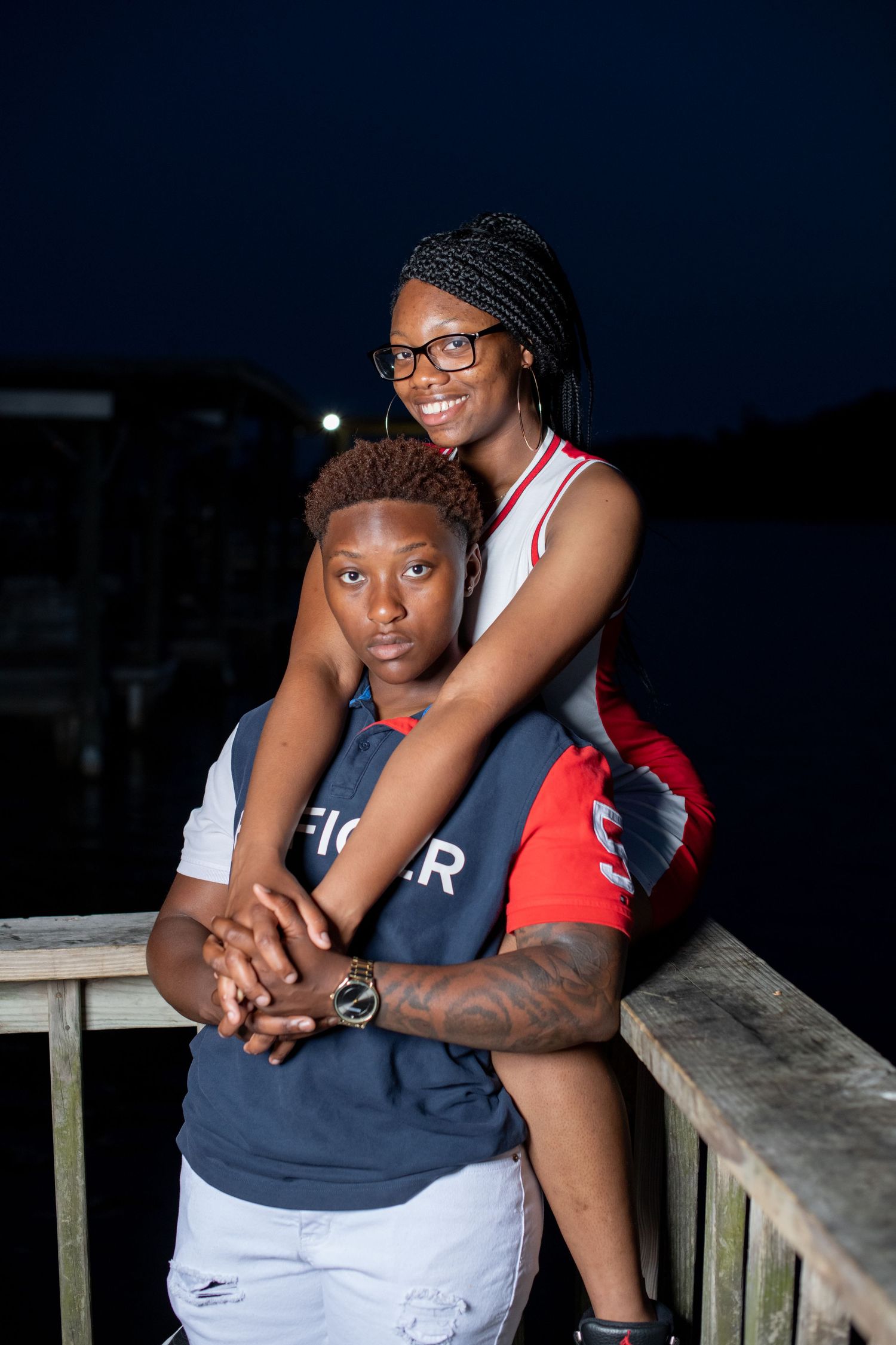 Couple posing at night on a wooden dock, woman embracing partner from behind near the water.