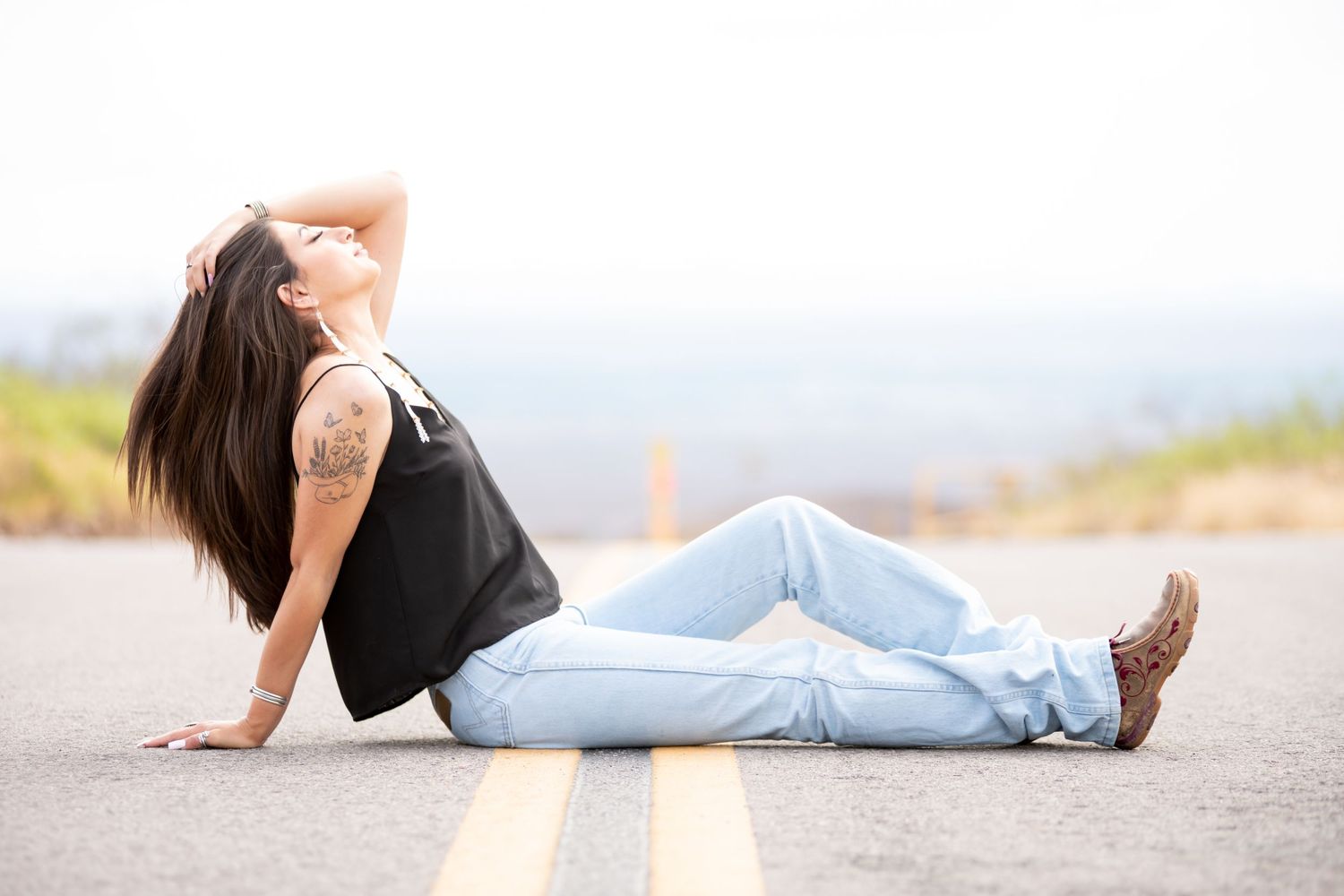 Woman with long dark hair and tattoo sitting on road in jeans and black tank top.