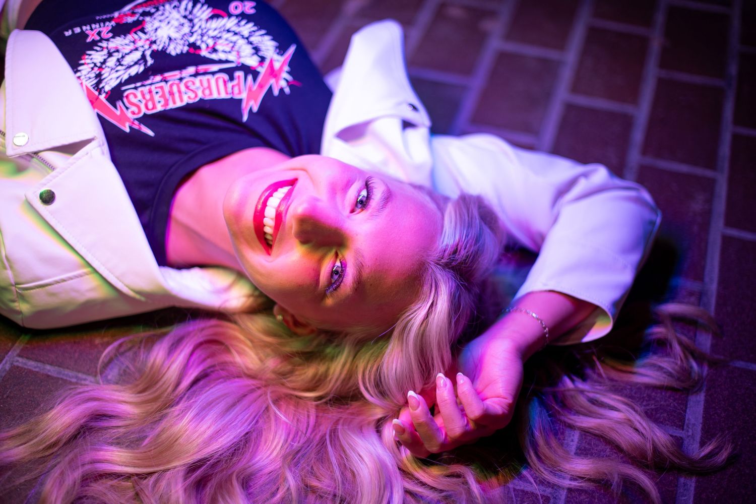 Smiling blonde woman lying on ground under purple neon lights wearing graphic tee and white jacket.