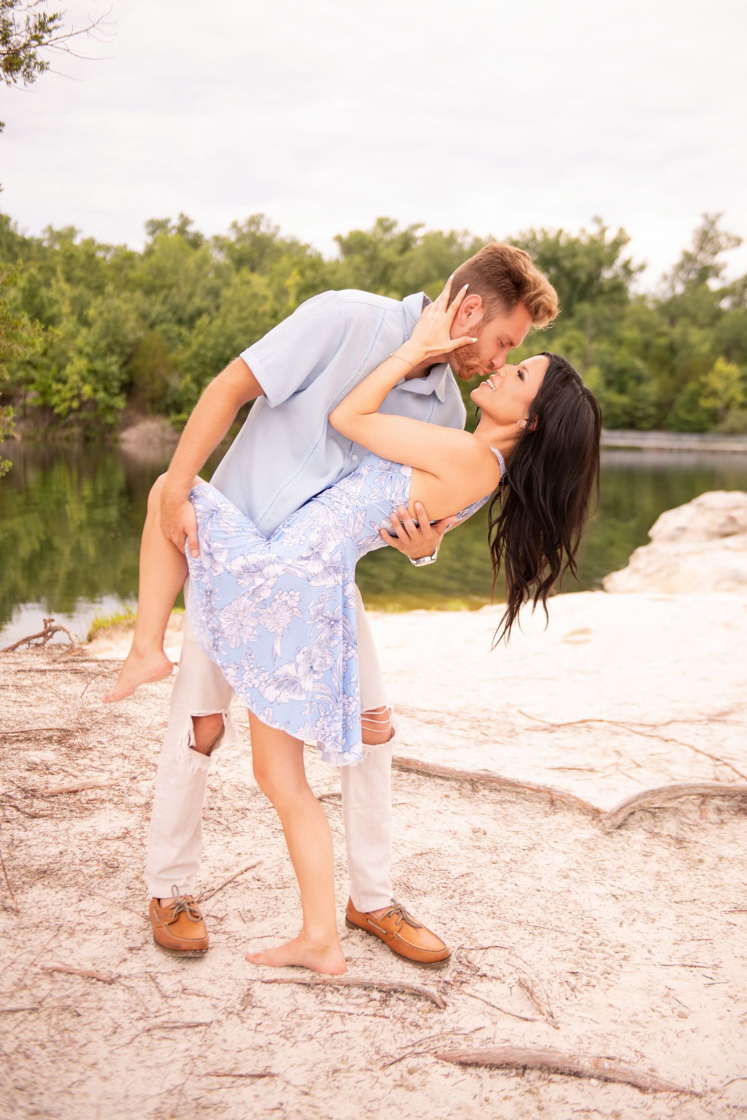 Man lifting woman in blue floral dress for a kiss on rocky terrain with lake and green trees in background.