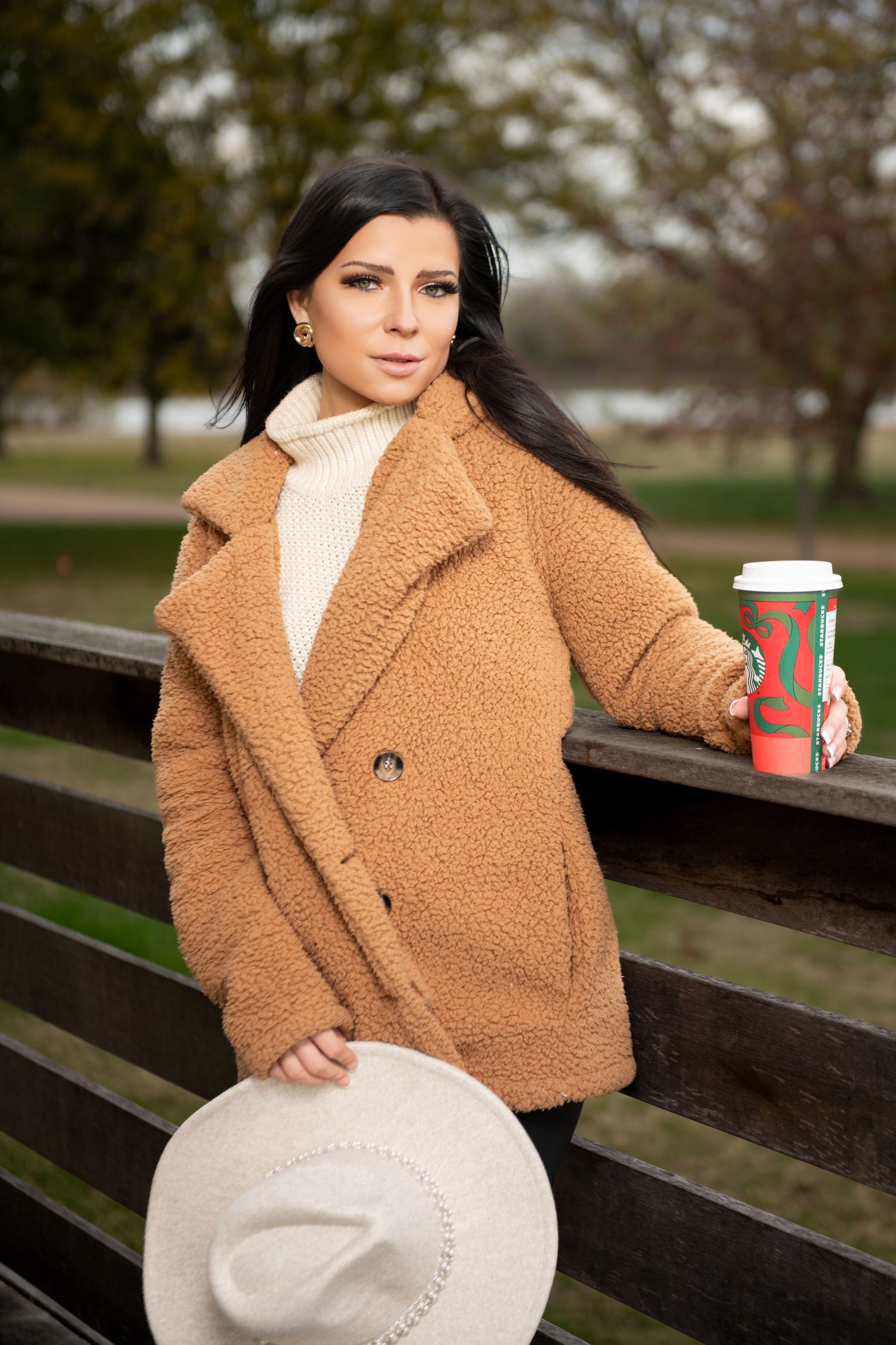 Dark-haired woman in camel coat holding white hat and red coffee cup outdoors in park.
