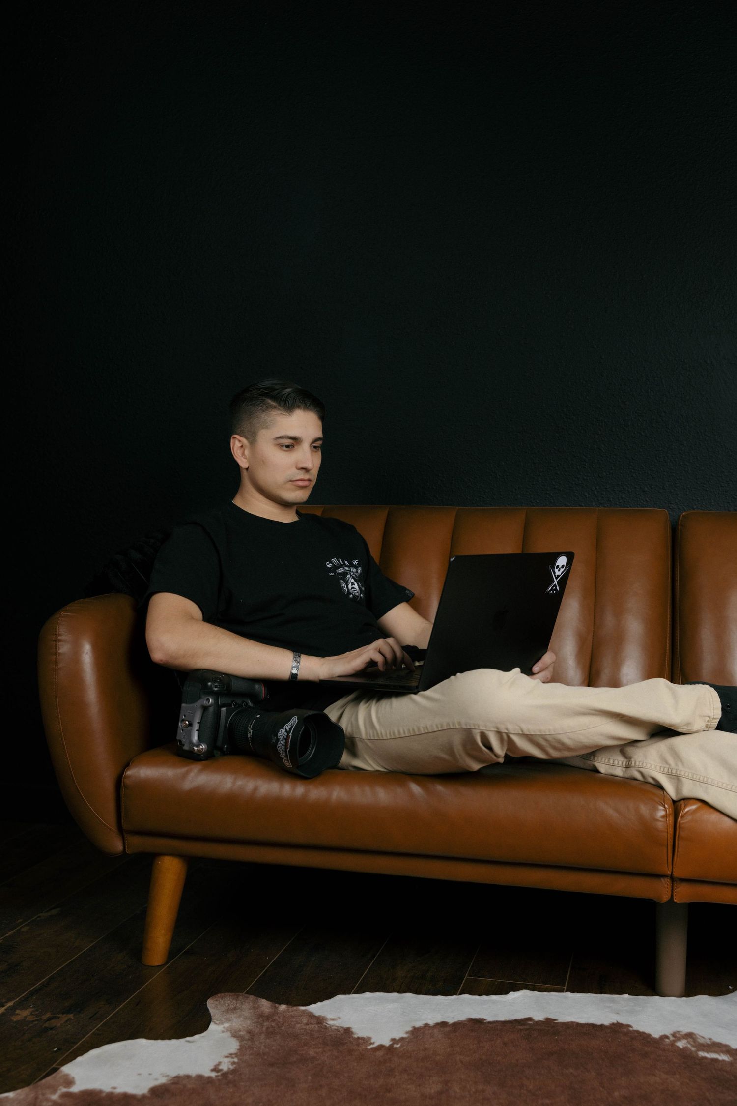 Man in black t-shirt working on laptop while sitting on brown leather sofa with camera nearby.