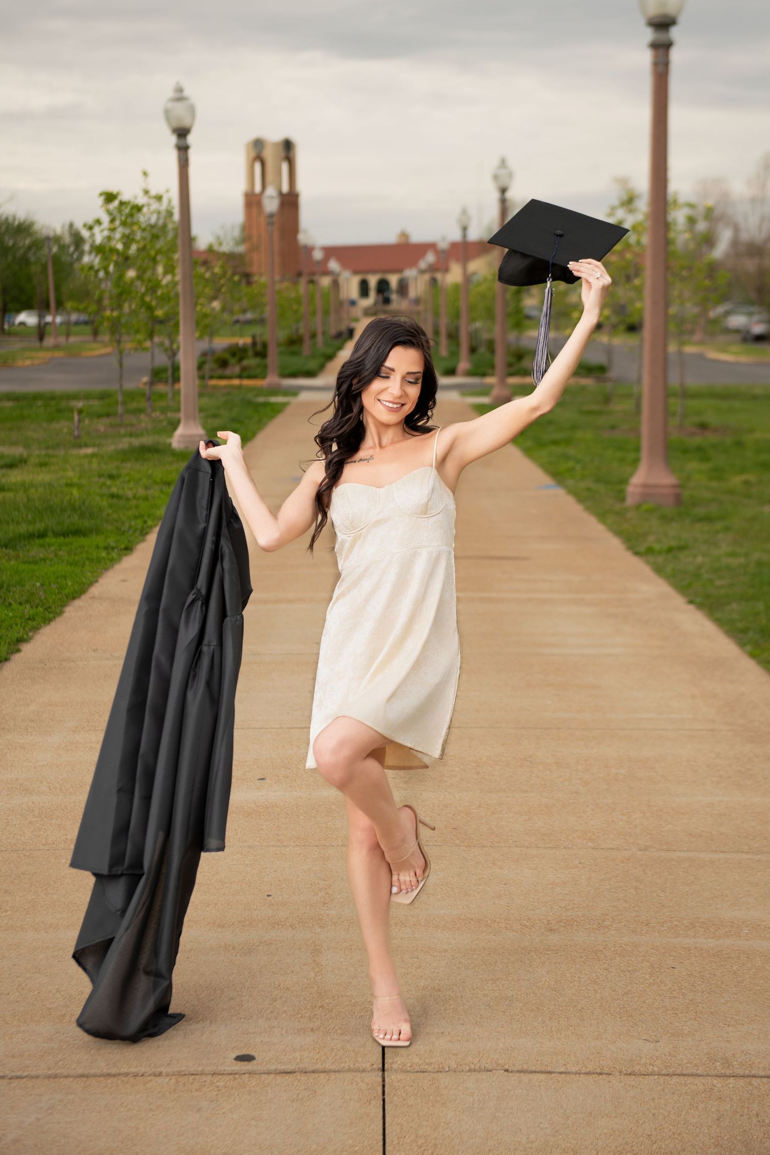 Joyful graduate in white dress celebrating outdoors holding cap and gown on campus pathway.