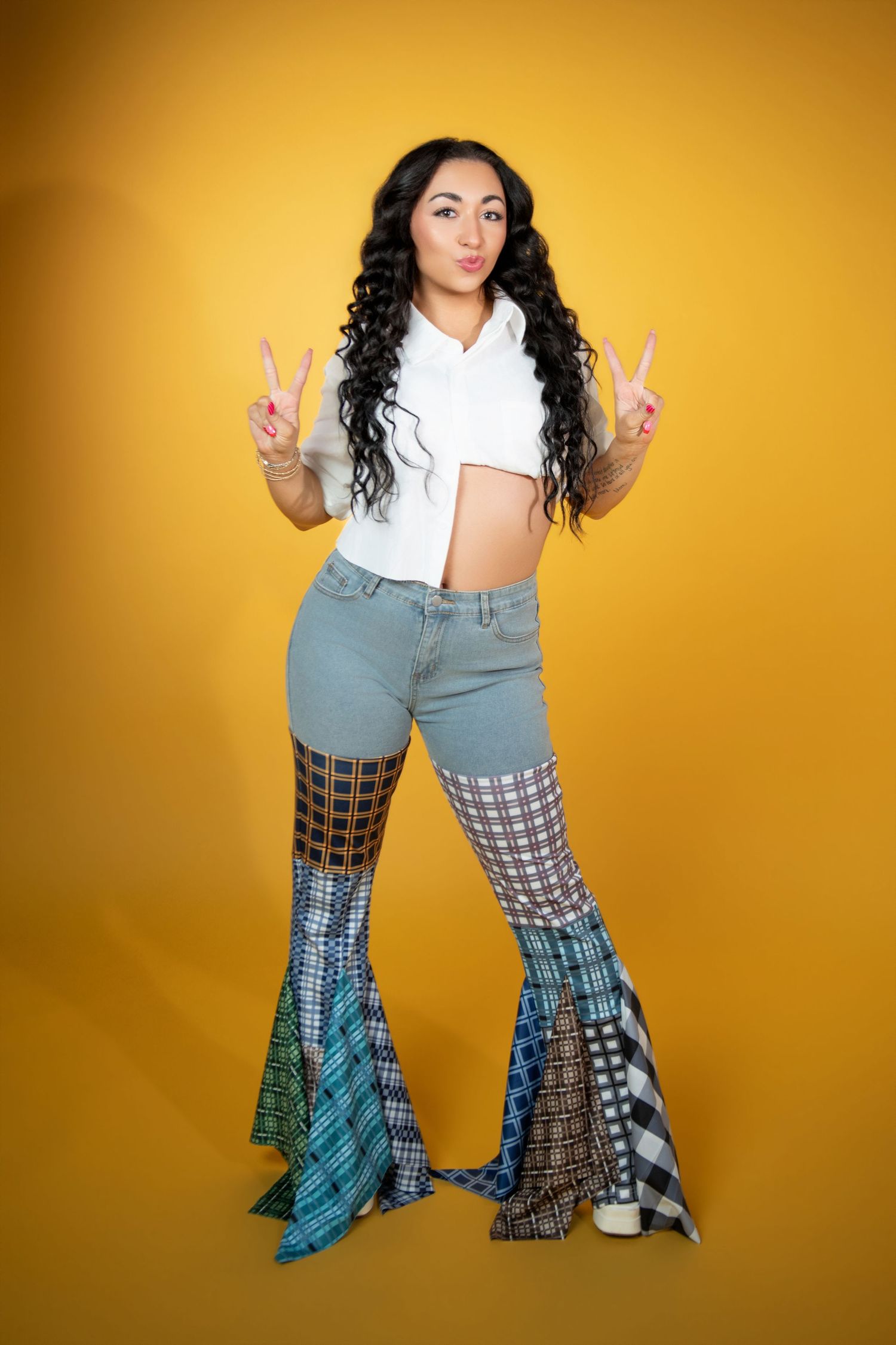 Young woman with curly hair in white crop top and patchwork bell-bottom jeans on yellow background.
