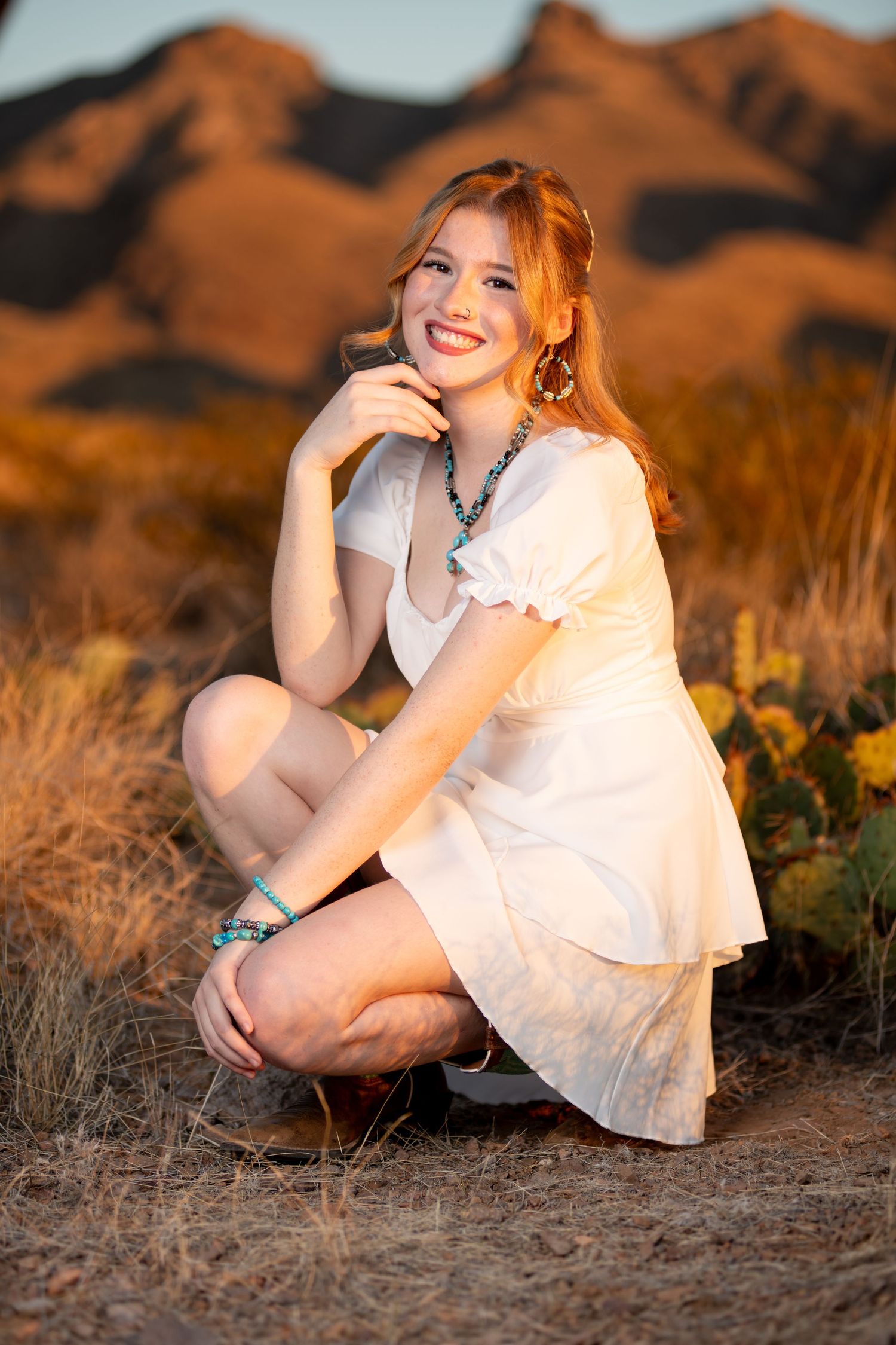 Smiling redhead woman in white dress crouching in desert landscape with golden mountain backdrop.