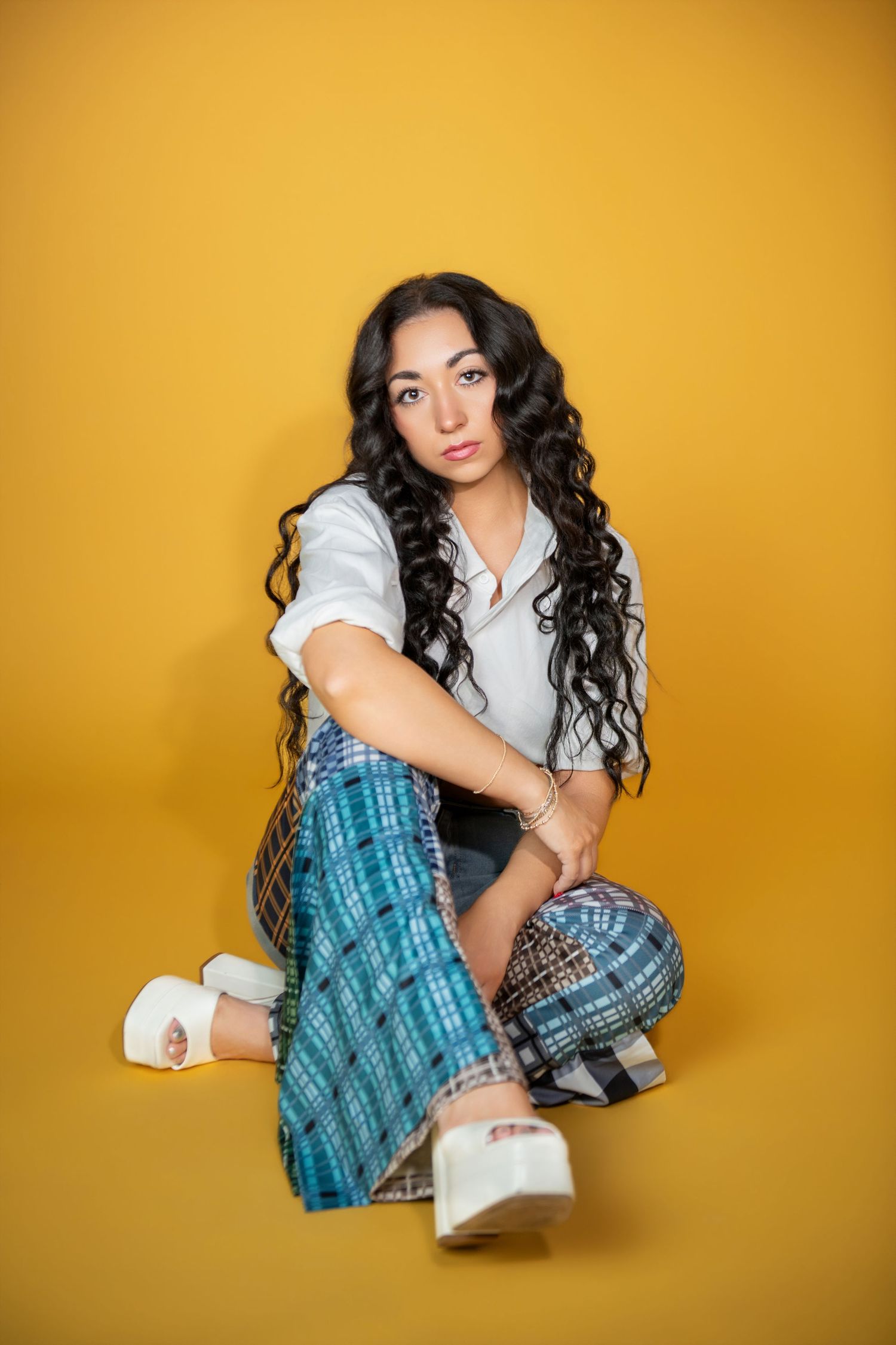 Brunette woman with curly hair sitting cross-legged in casual outfit on yellow background.
