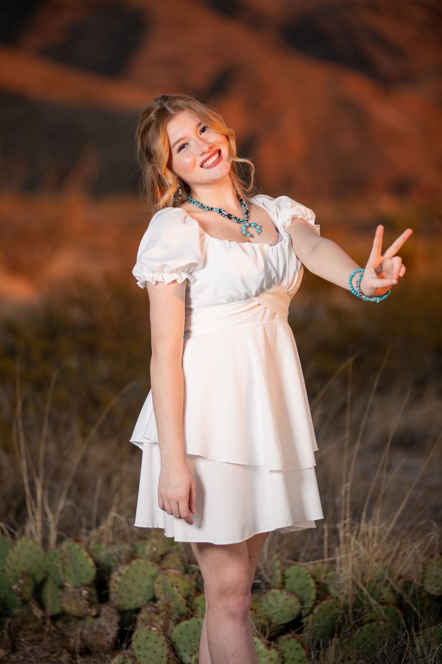 Smiling blonde woman in white dress flashing peace sign near cacti with red mountains behind her.