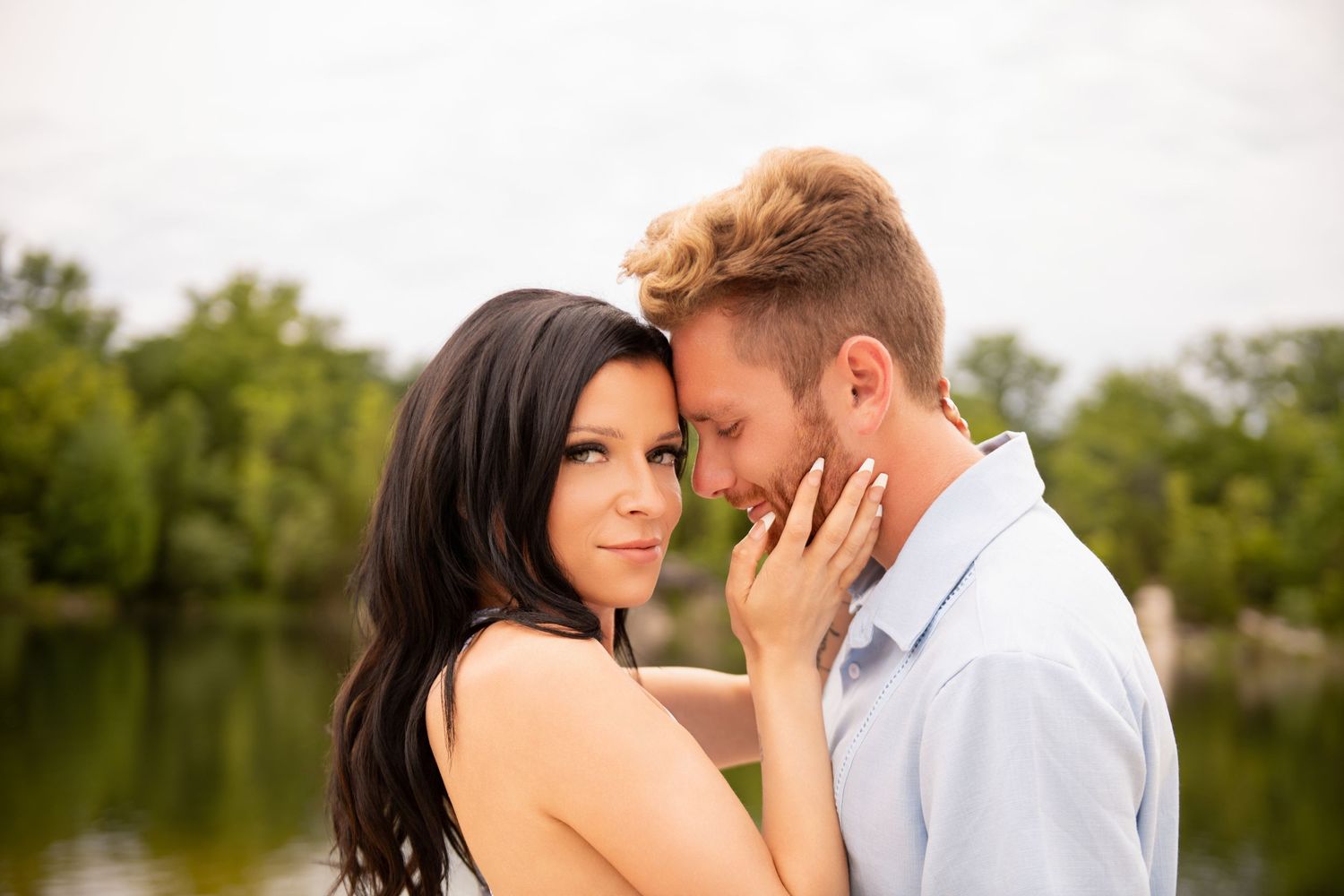 Romantic couple sharing an intimate moment near a peaceful lake surrounded by green trees.