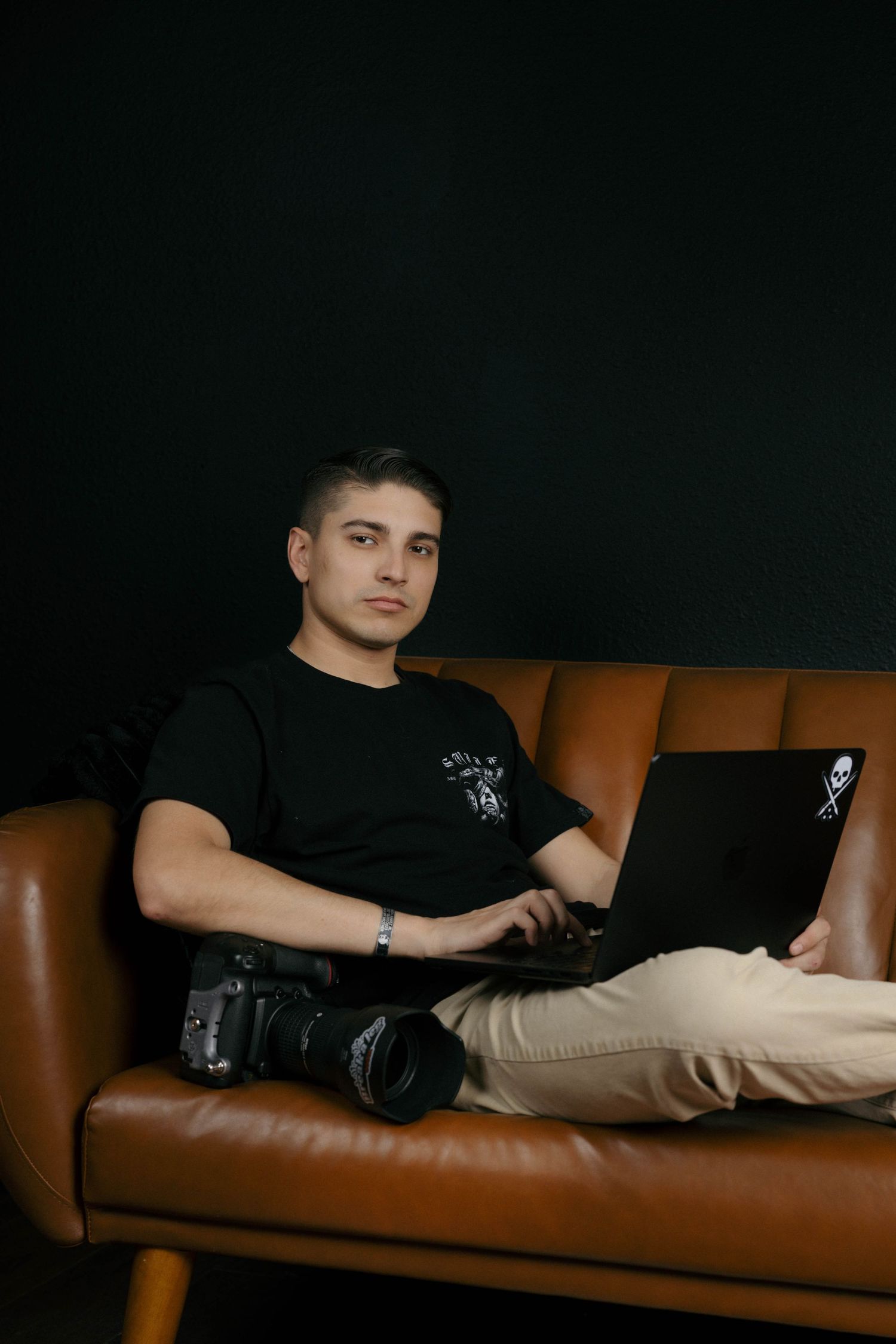 Young man in black t-shirt working on laptop on brown leather sofa with camera nearby.