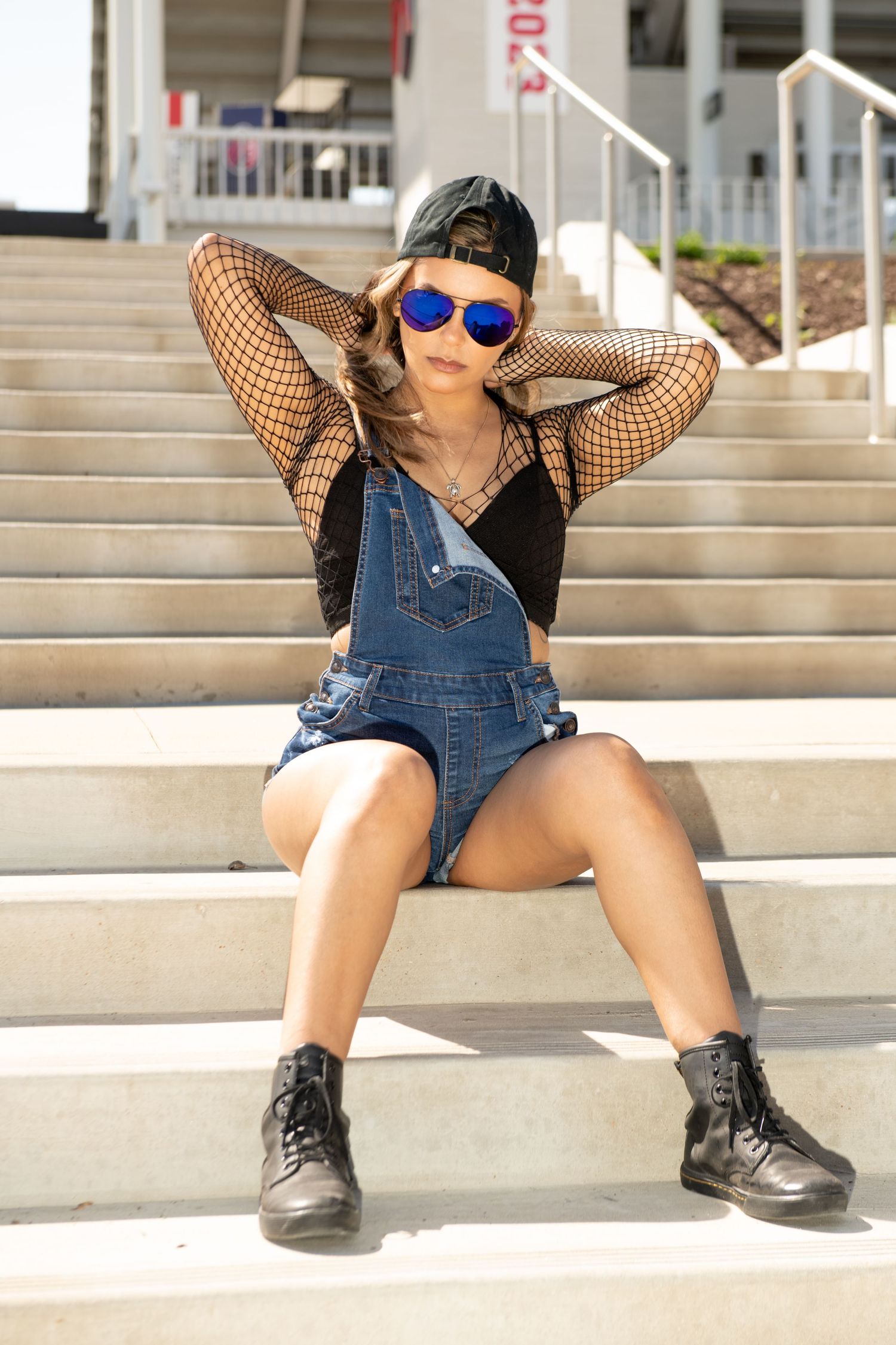 Young woman in denim overalls, fishnet top, cap, and blue mirrored sunglasses sitting on outdoor steps.