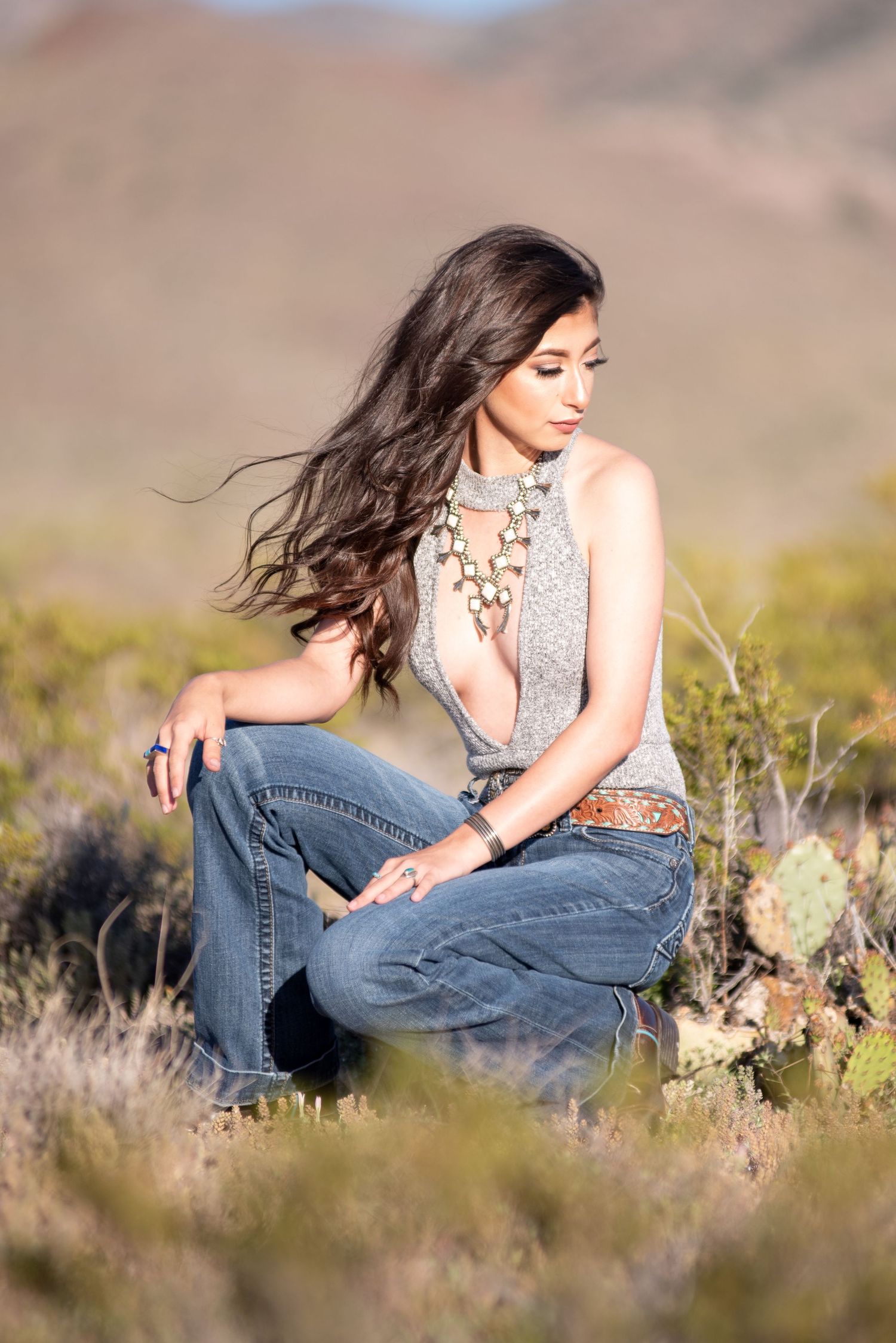 Woman in gray halter top and flared jeans crouching in desert landscape.