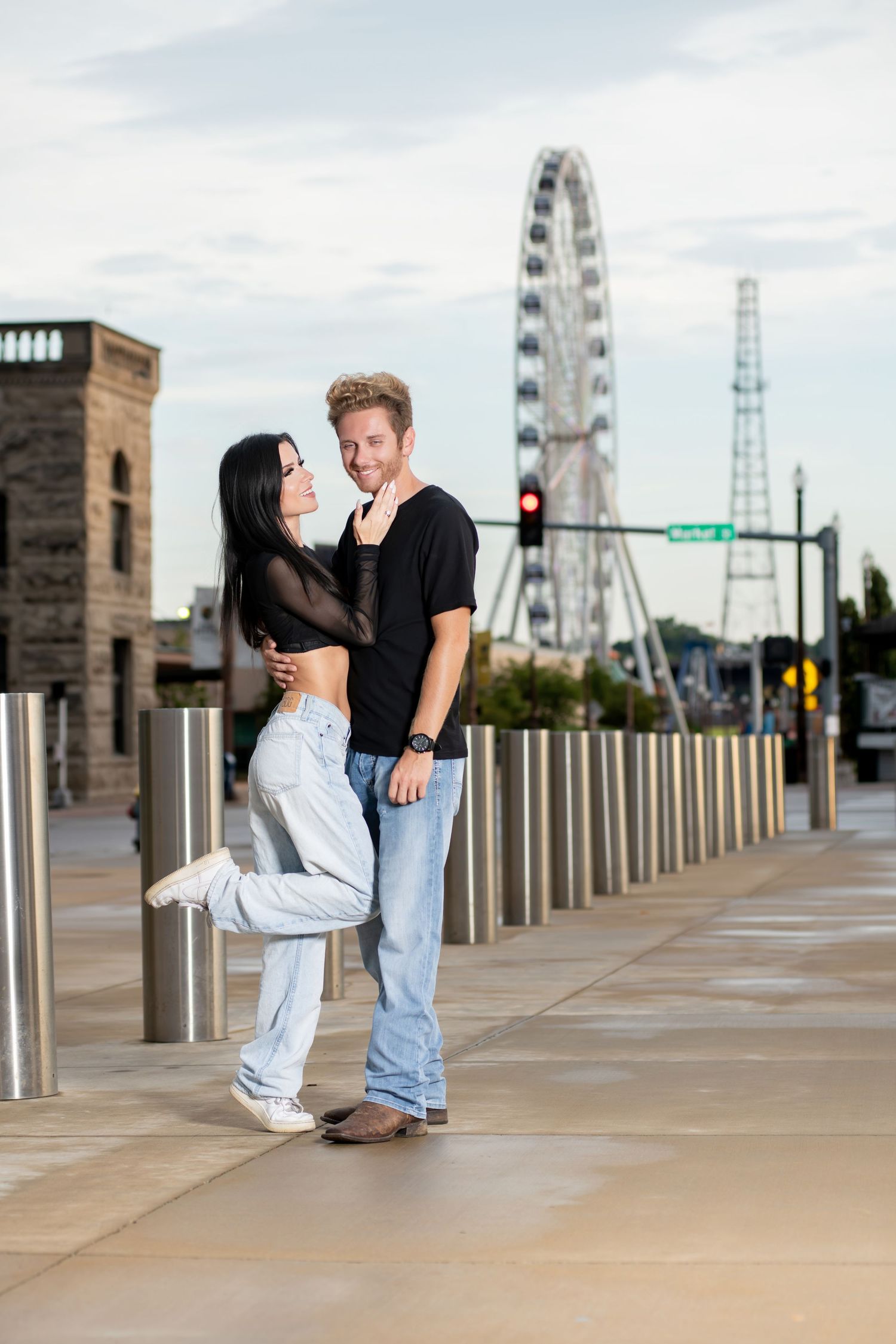 Couple embracing on urban walkway with Ferris wheel in background, woman in black top and jeans, man in black shirt.