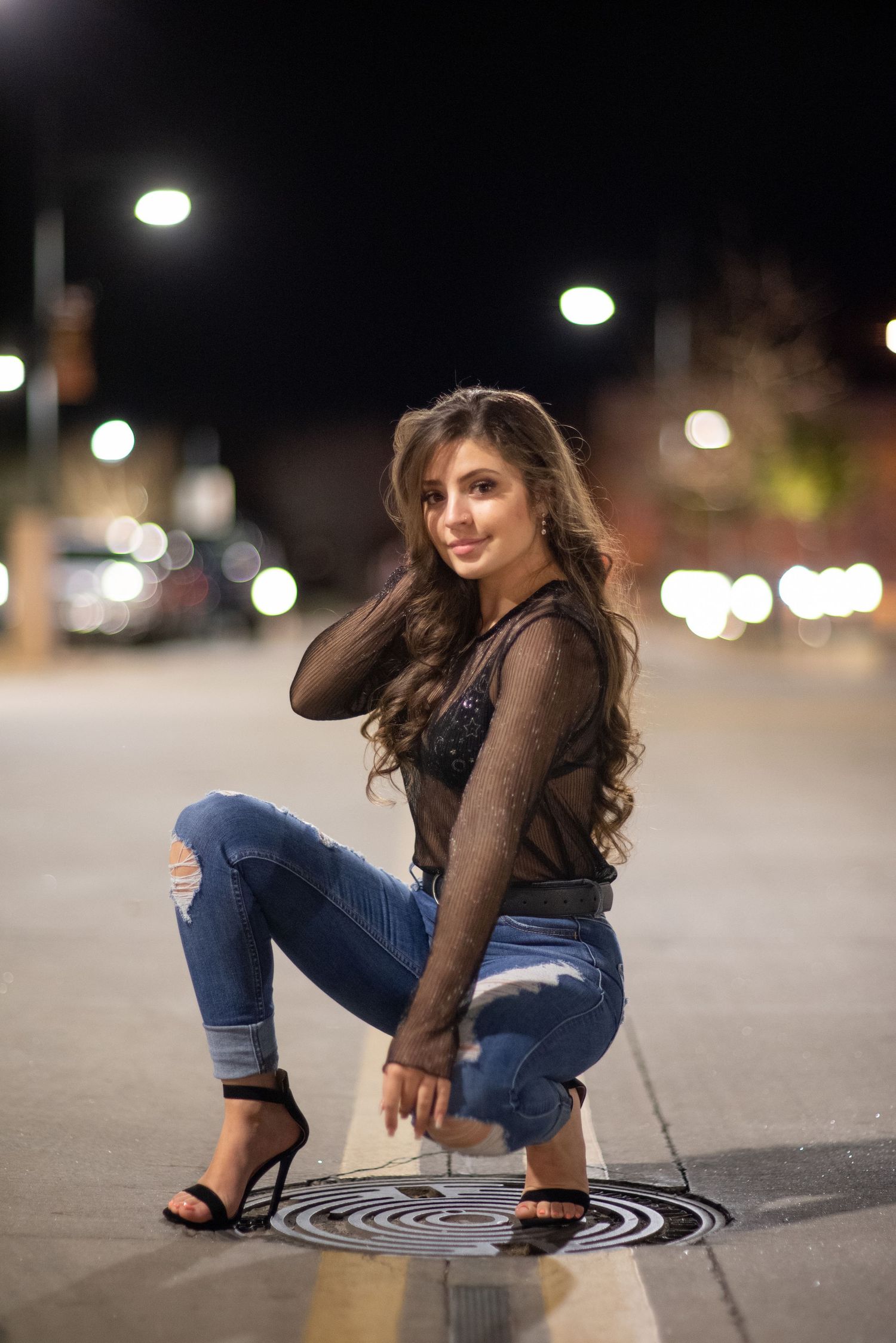 Woman in sheer black top and ripped jeans posing on urban street at night.