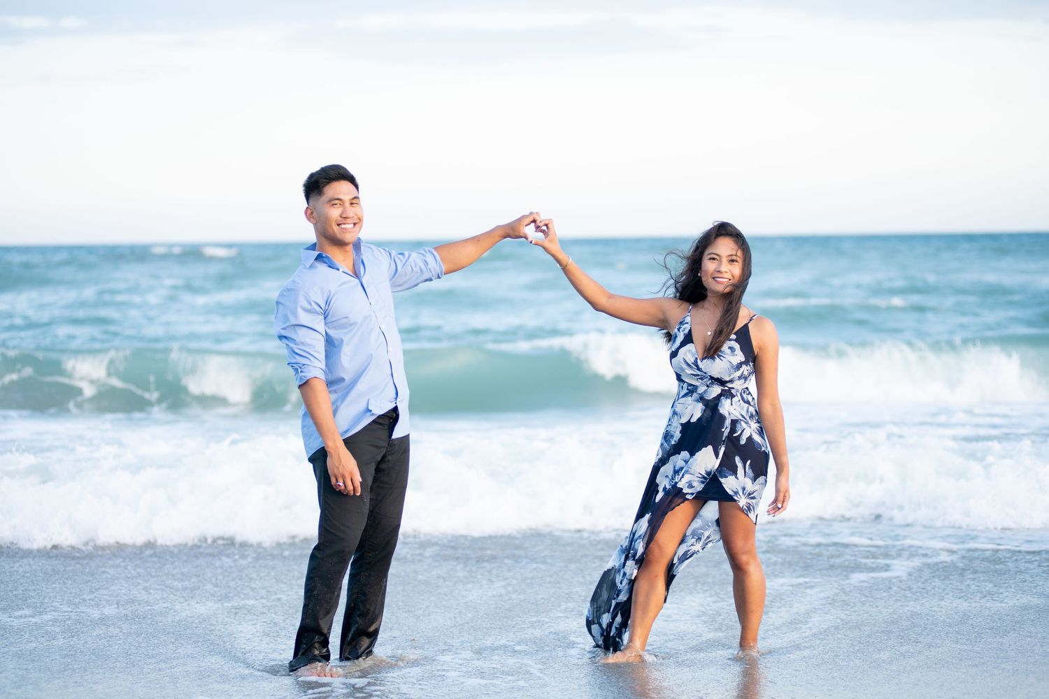 Couple dancing playfully in ocean waves, man in blue shirt and woman in floral maxi dress.