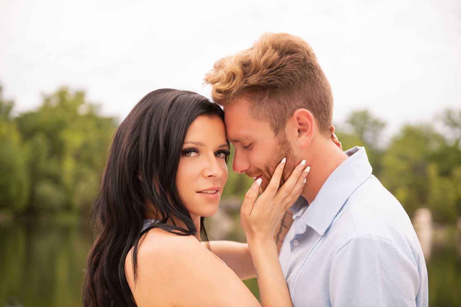 Intimate couple moment with foreheads touching near a lake with green trees in soft focus background.