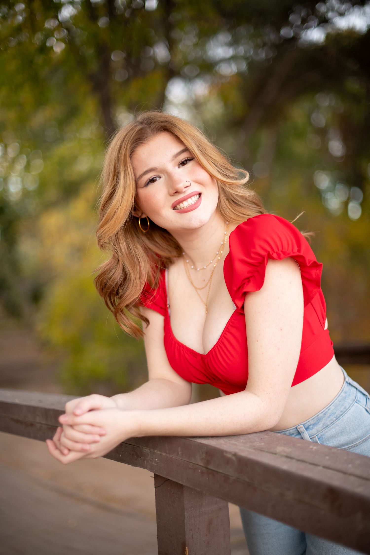 Smiling redhead woman in red crop top leaning on wooden railing surrounded by autumn foliage.