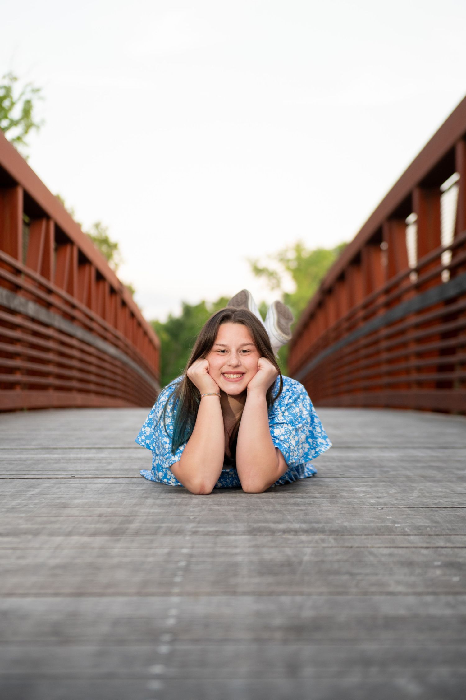 Smiling teenage girl lying on wooden bridge deck with red railings in blue floral dress.