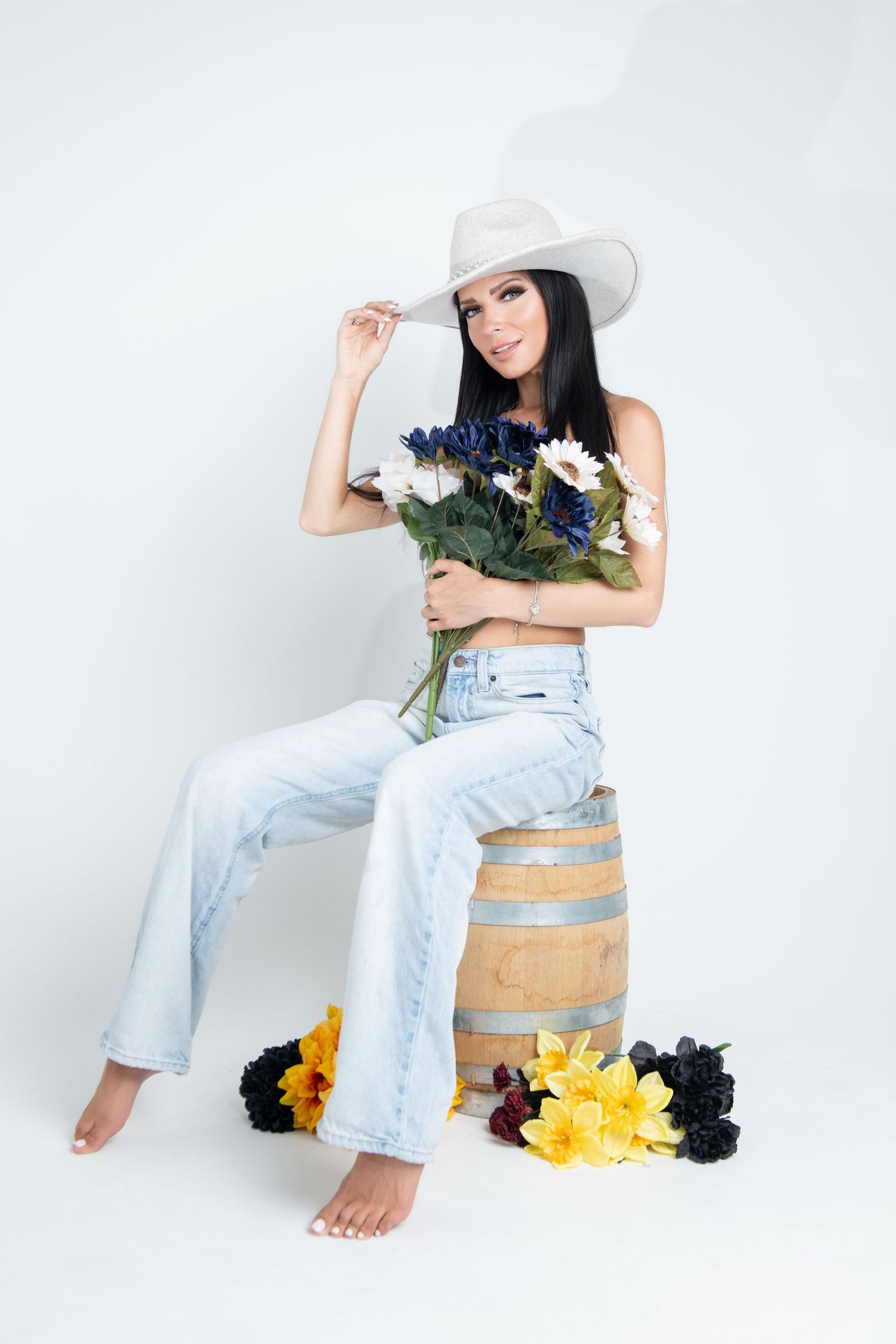 Woman in white cowboy hat and jeans sitting on barrel with flowers.