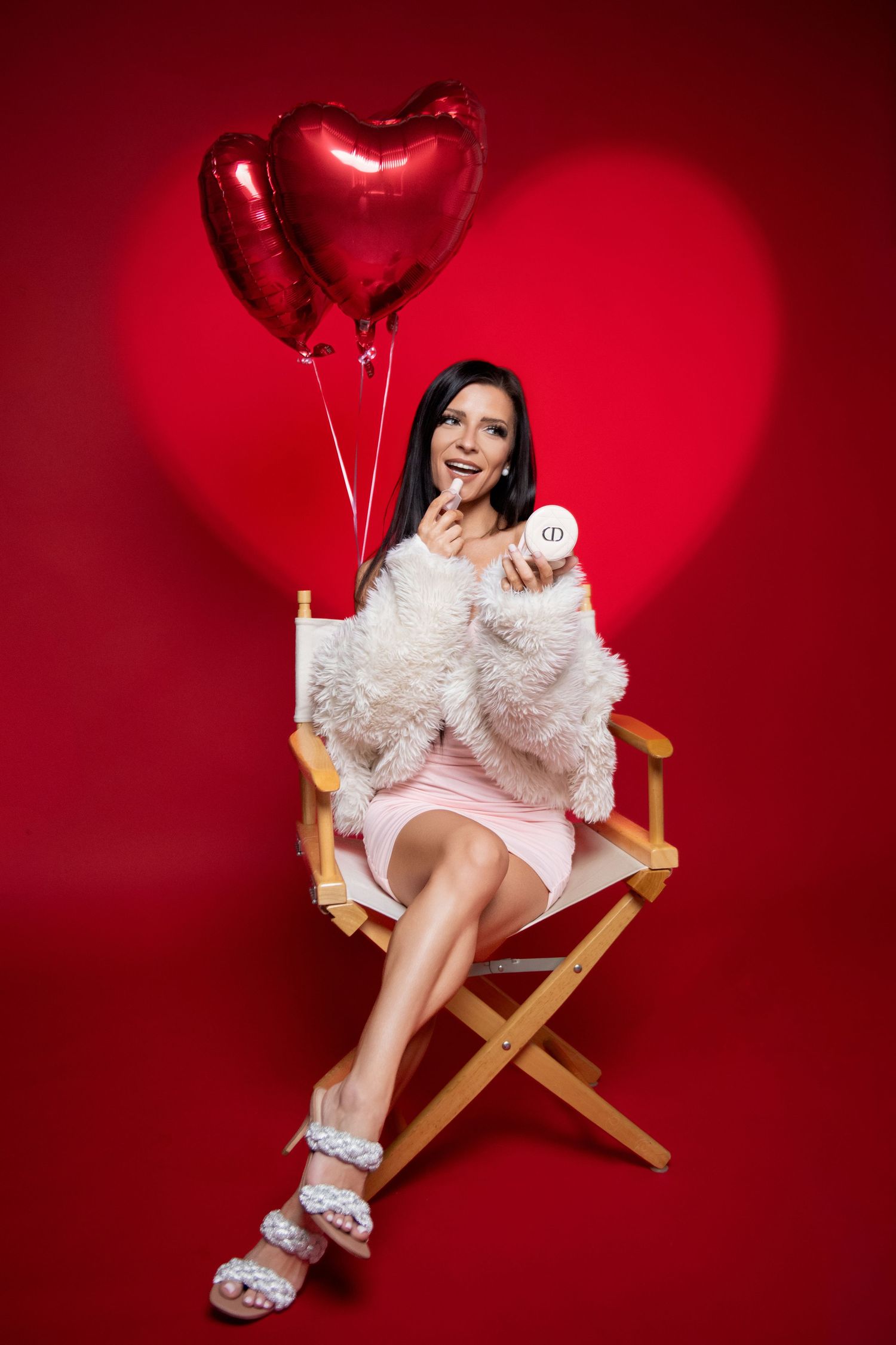 Smiling woman in pink dress holding heart balloons seated on chair against red background.
