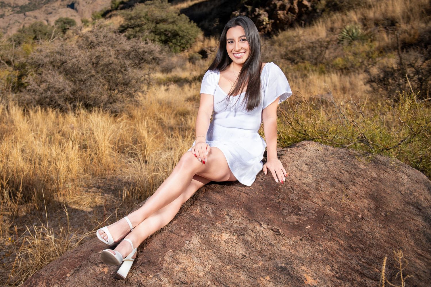 Smiling young woman in white dress seated on a boulder in a natural outdoor setting.