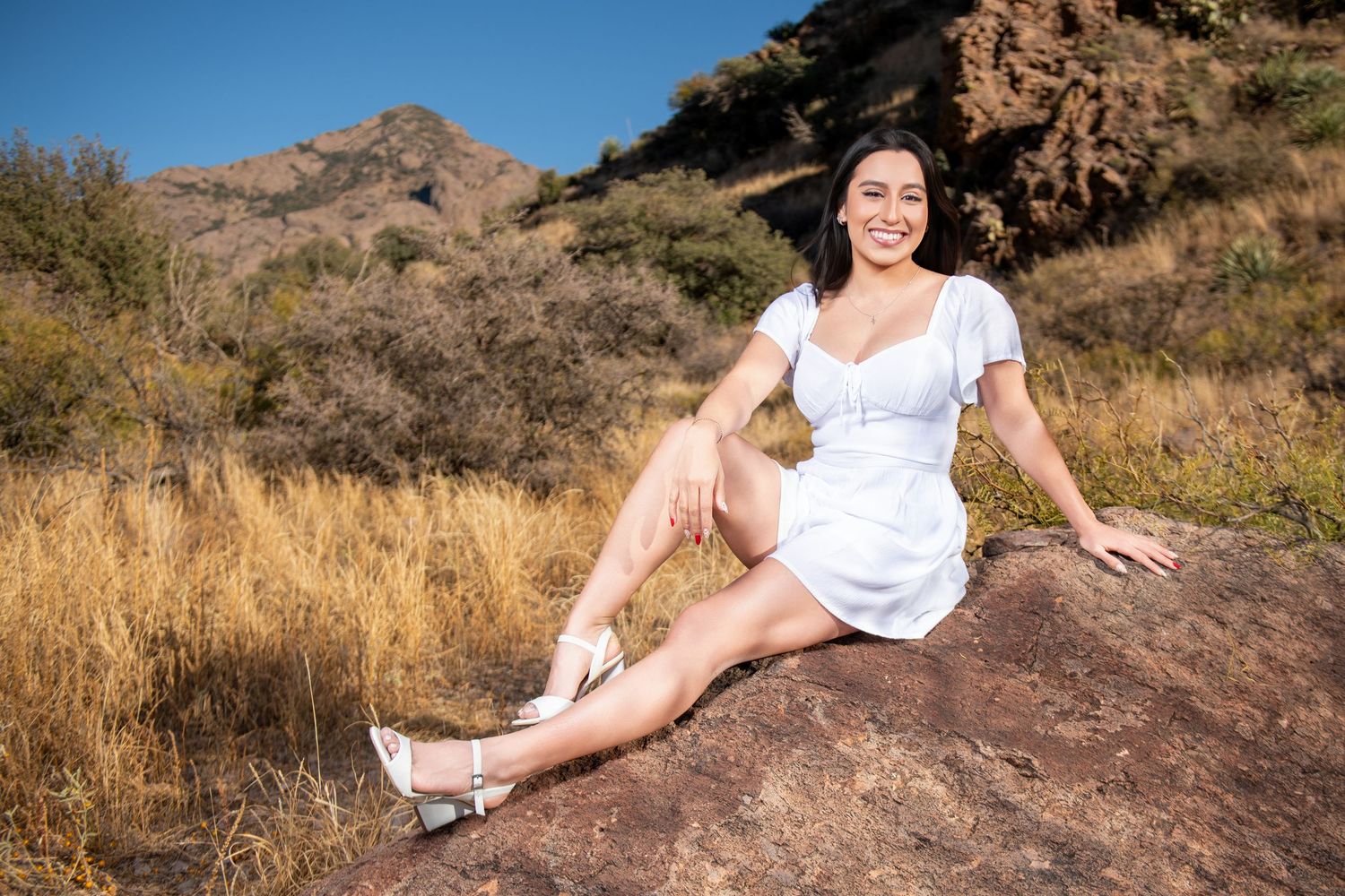 Smiling woman in white dress sits on desert rock with mountain landscape behind her.
