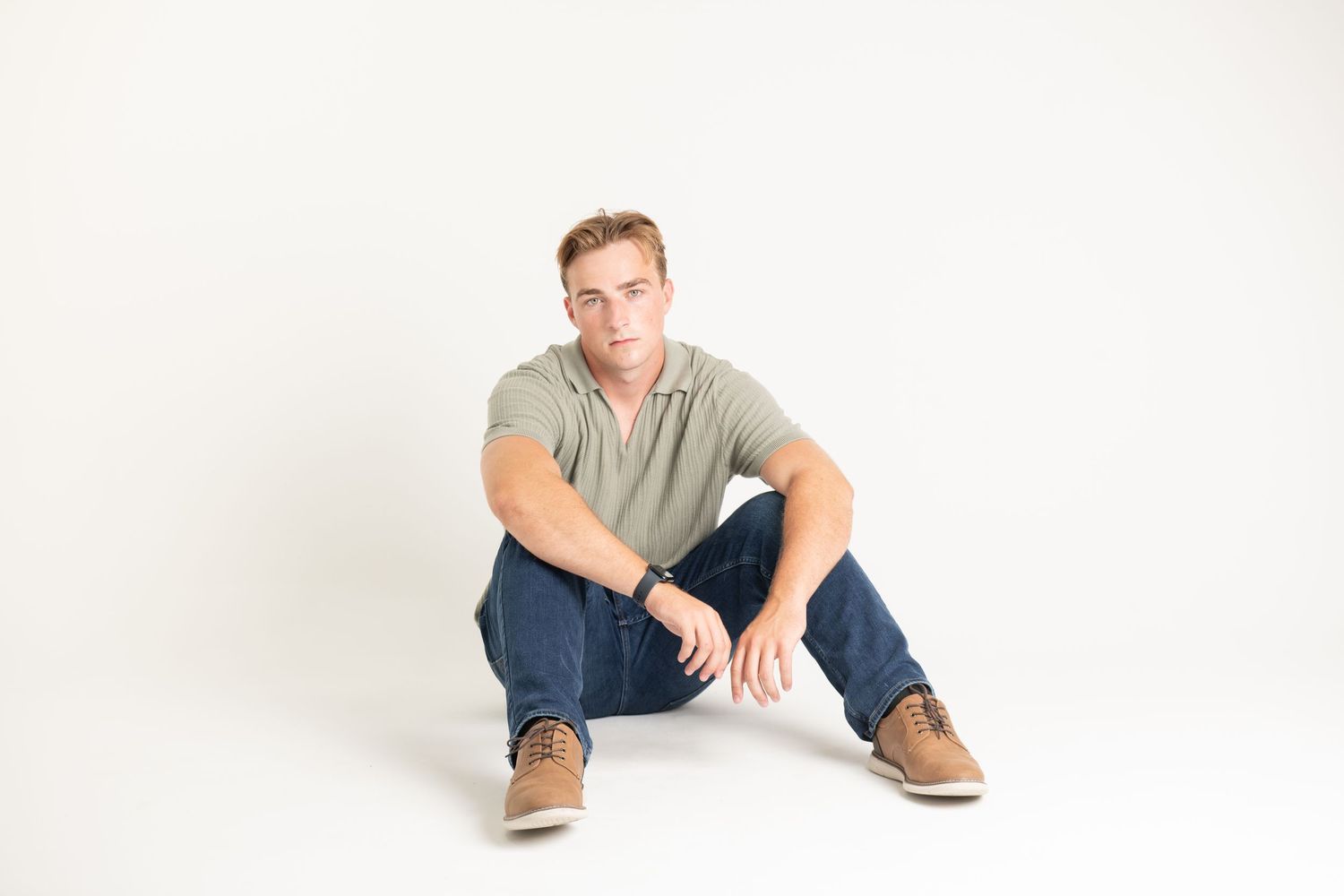 Young man in sage green polo and jeans poses seated on white studio floor.