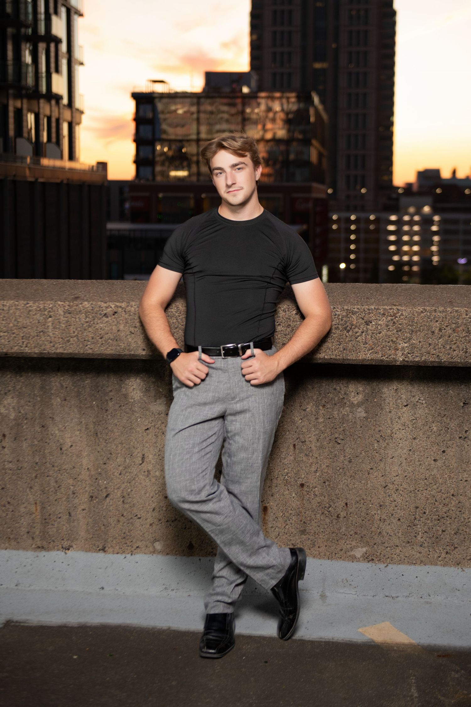 Muscular young man in black tee and gray pants posing on rooftop with city sunset skyline.