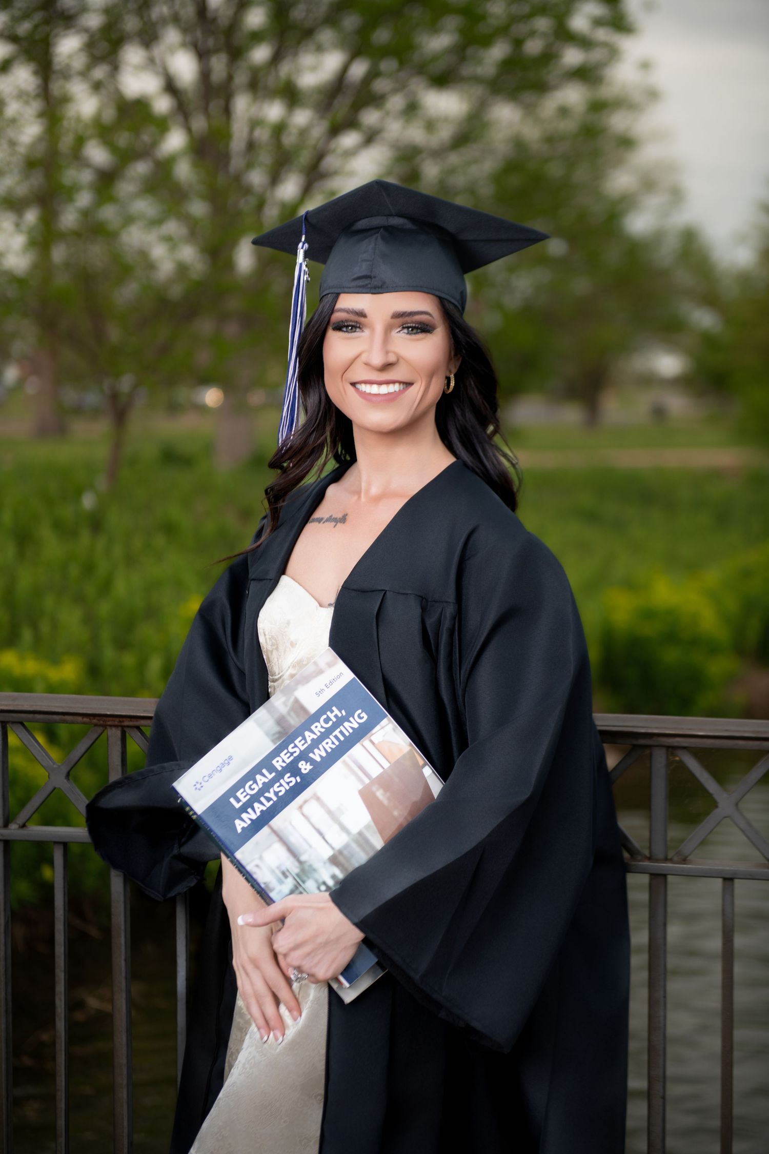 Smiling graduate in black cap and gown holding a Legal Research Analysis and Writing textbook outdoors.
