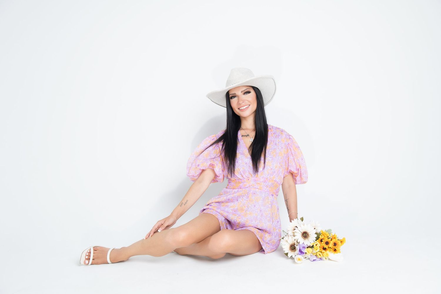 Smiling woman in pink dress and white hat sitting with flowers.