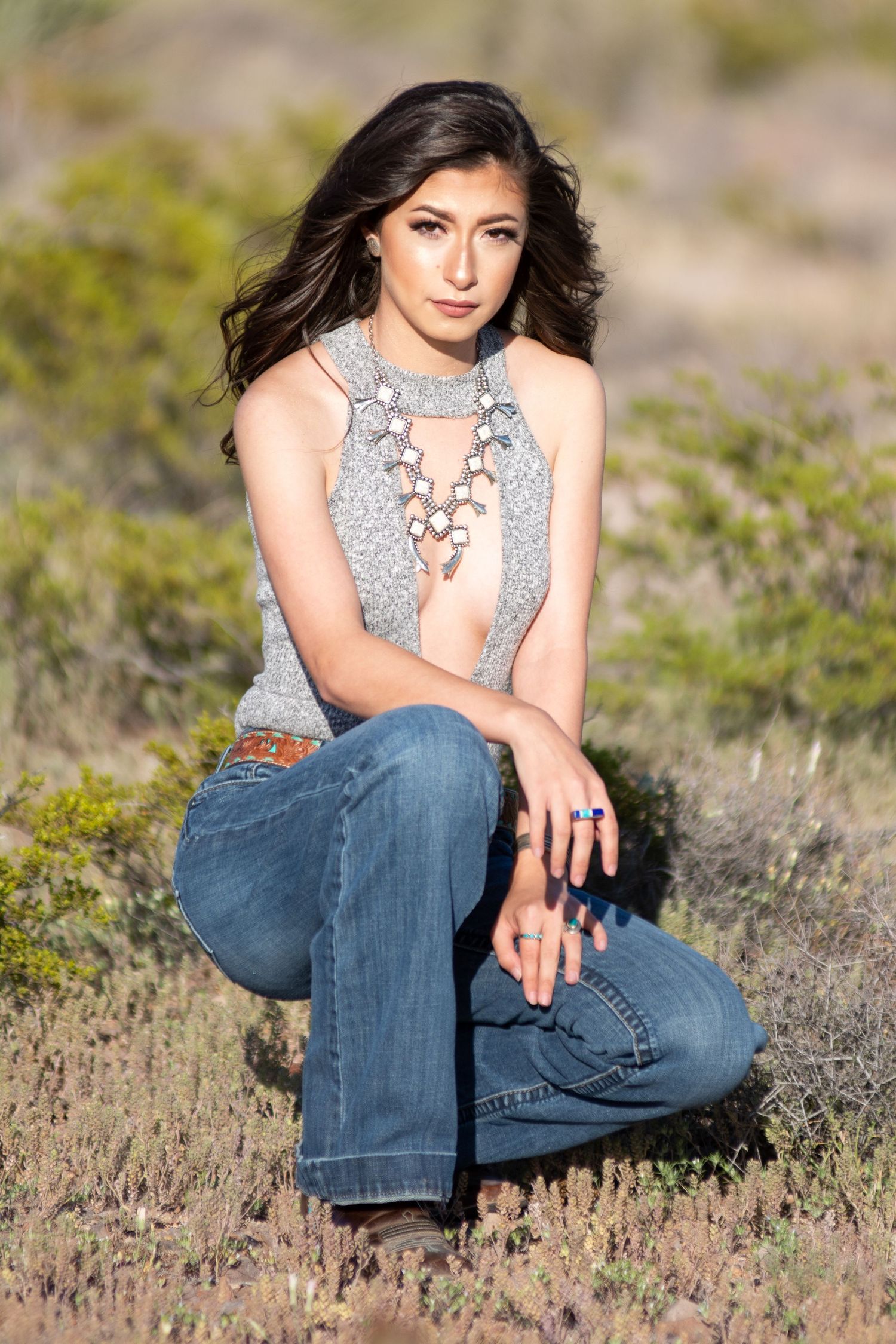Woman wearing silver squash blossom necklace and gray halter top crouching outdoors.