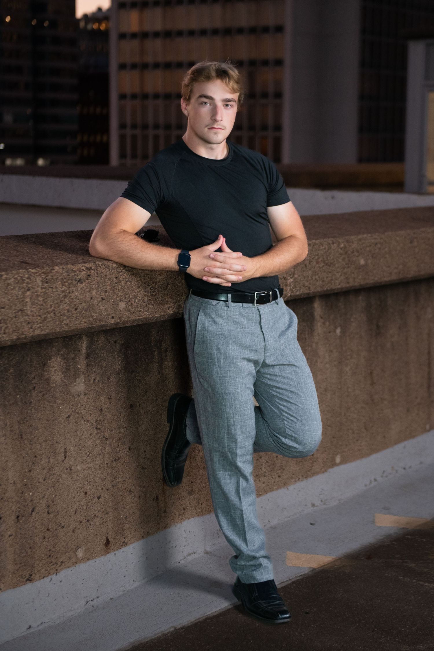 Athletic young man in black t-shirt and gray trousers leaning against concrete wall at dusk.