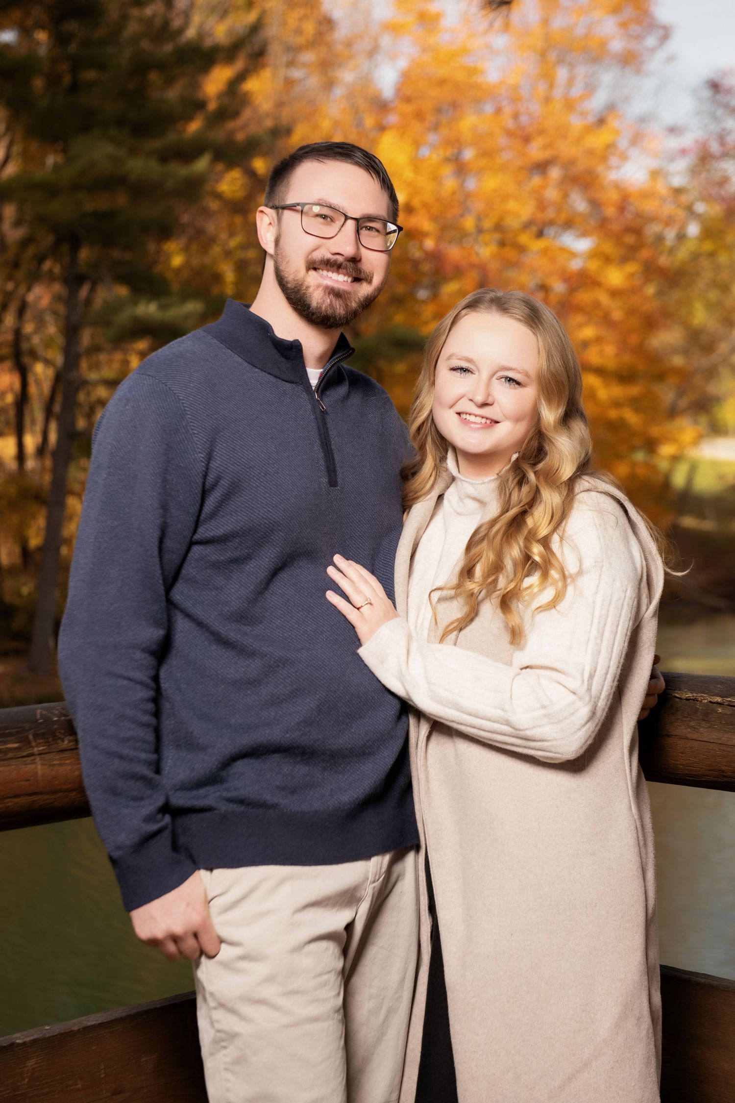 Smiling couple posing on bridge surrounded by vibrant autumn foliage, man in navy pullover, woman in cream sweater.