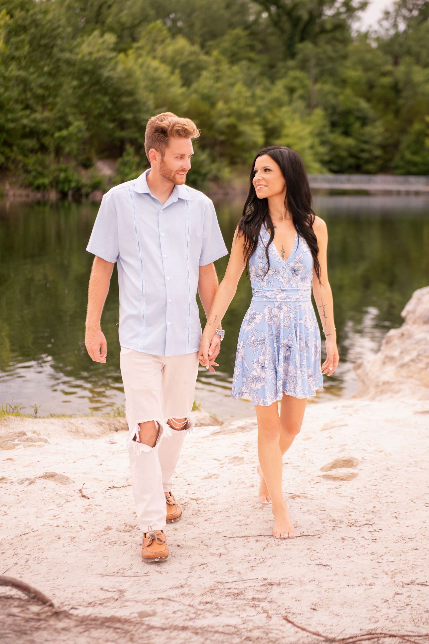 Happy couple holding hands and walking along rocky lakeside path, woman in blue floral dress, man in light blue shirt.