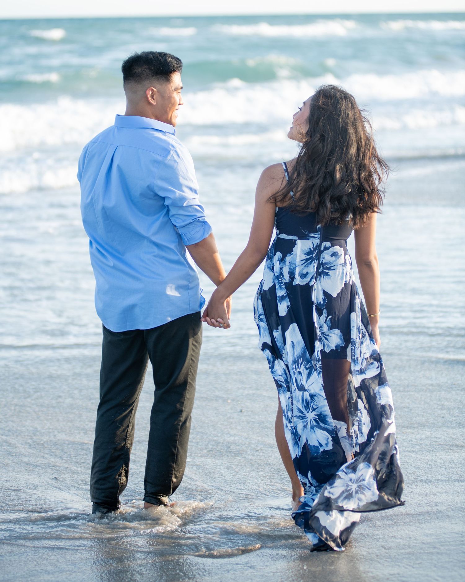 Couple holding hands walking along the shoreline, woman in flowing blue floral dress.