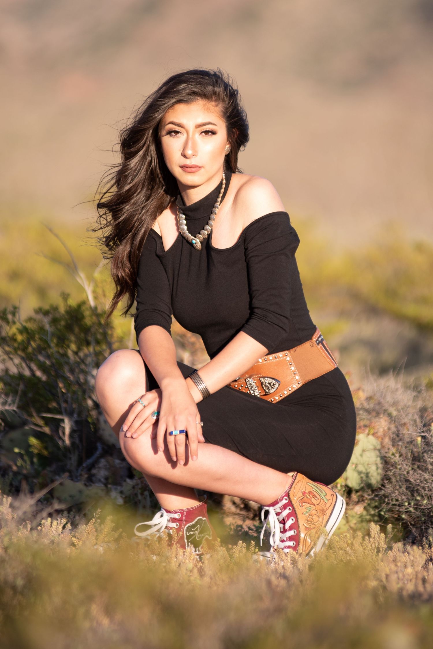 Woman in black off-shoulder dress and tooled leather belt posing in desert field.
