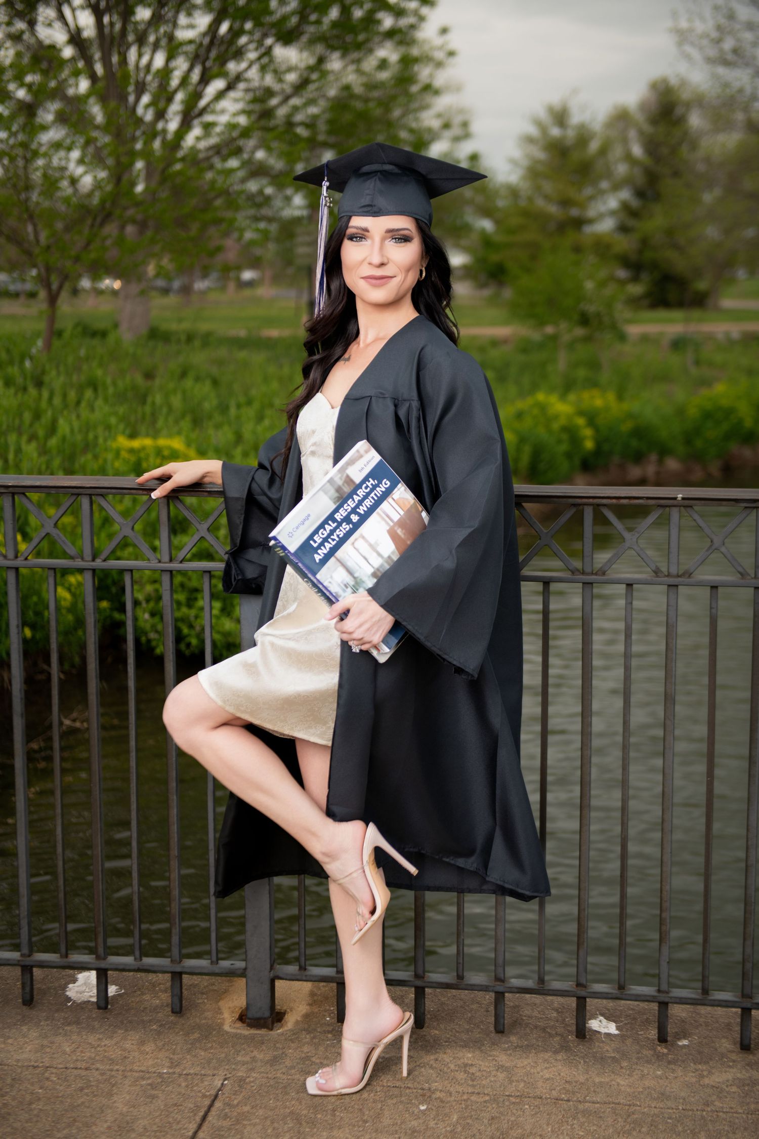 Brunette woman in graduation cap and gown leaning on iron fence holding textbook outdoors.
