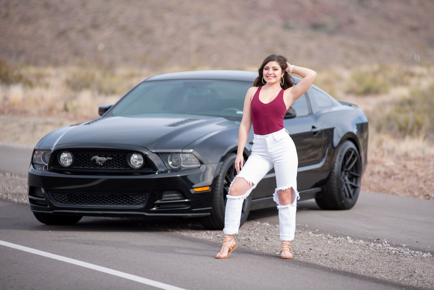 Woman in burgundy top and white jeans posing beside black Ford Mustang on road.
