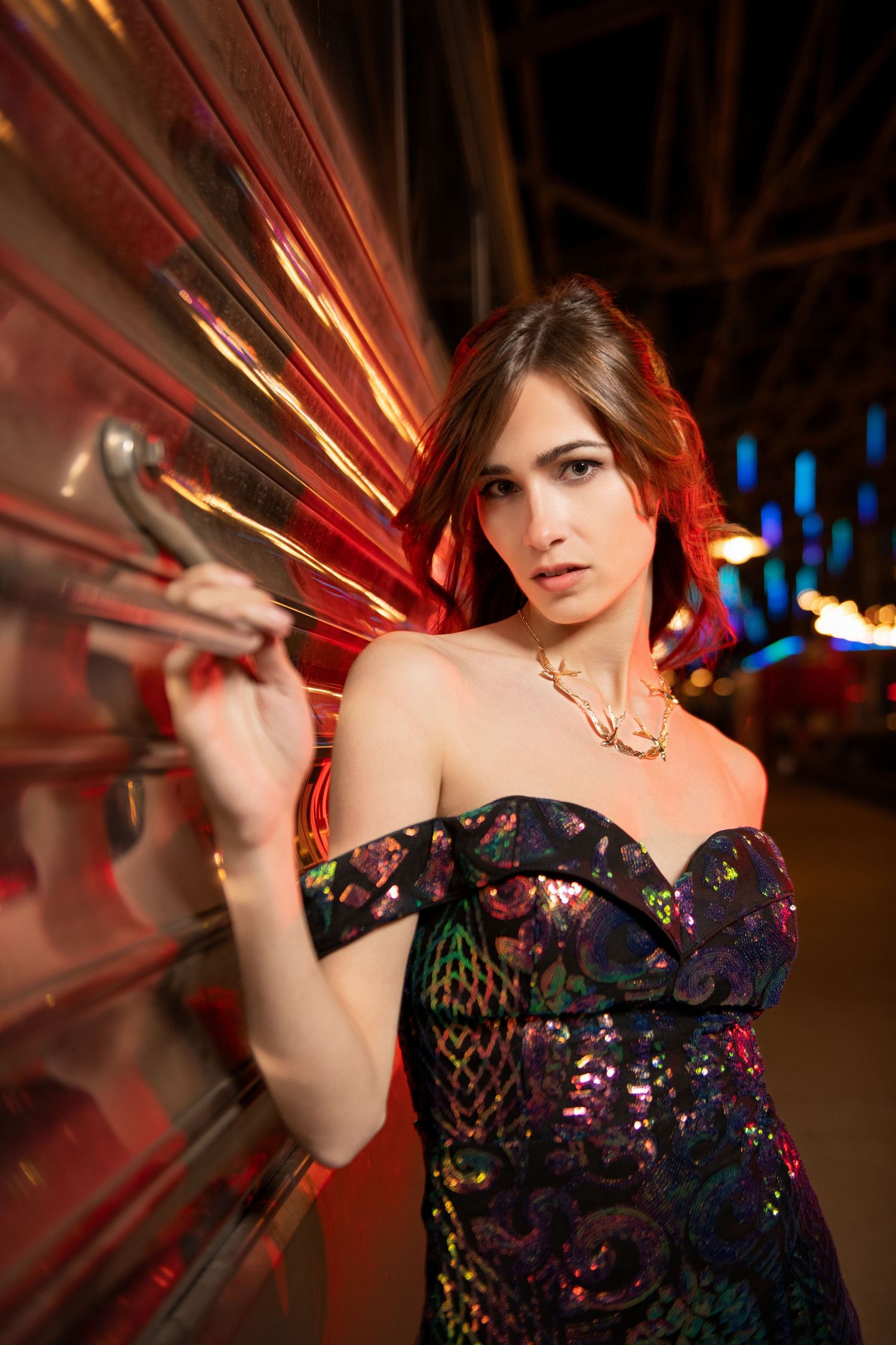 Woman in colorful sequin off-shoulder gown posing near illuminated carnival ride at night.