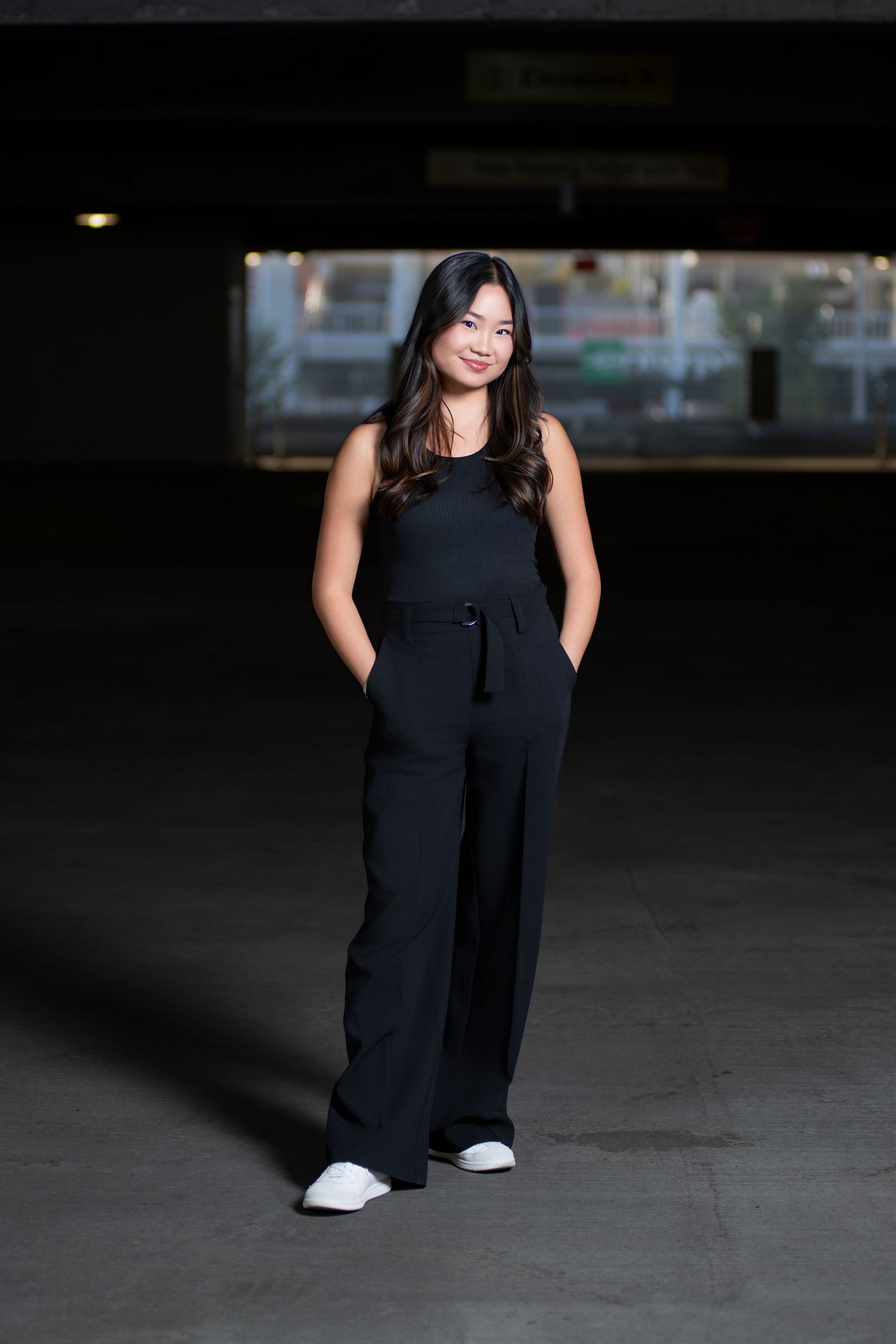 Young Asian woman in black sleeveless jumpsuit and white sneakers posing in dark urban parking structure.