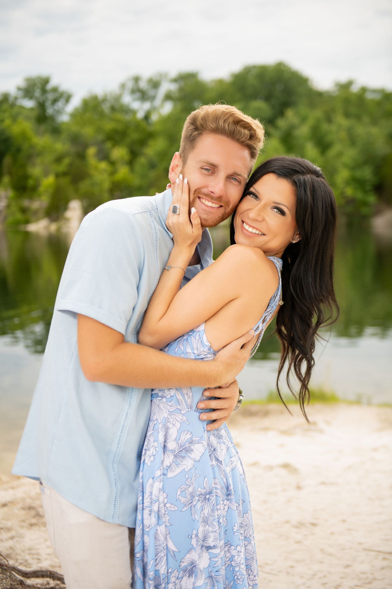 Happy couple embracing outdoors near a lake, woman in blue floral dress smiling at camera.