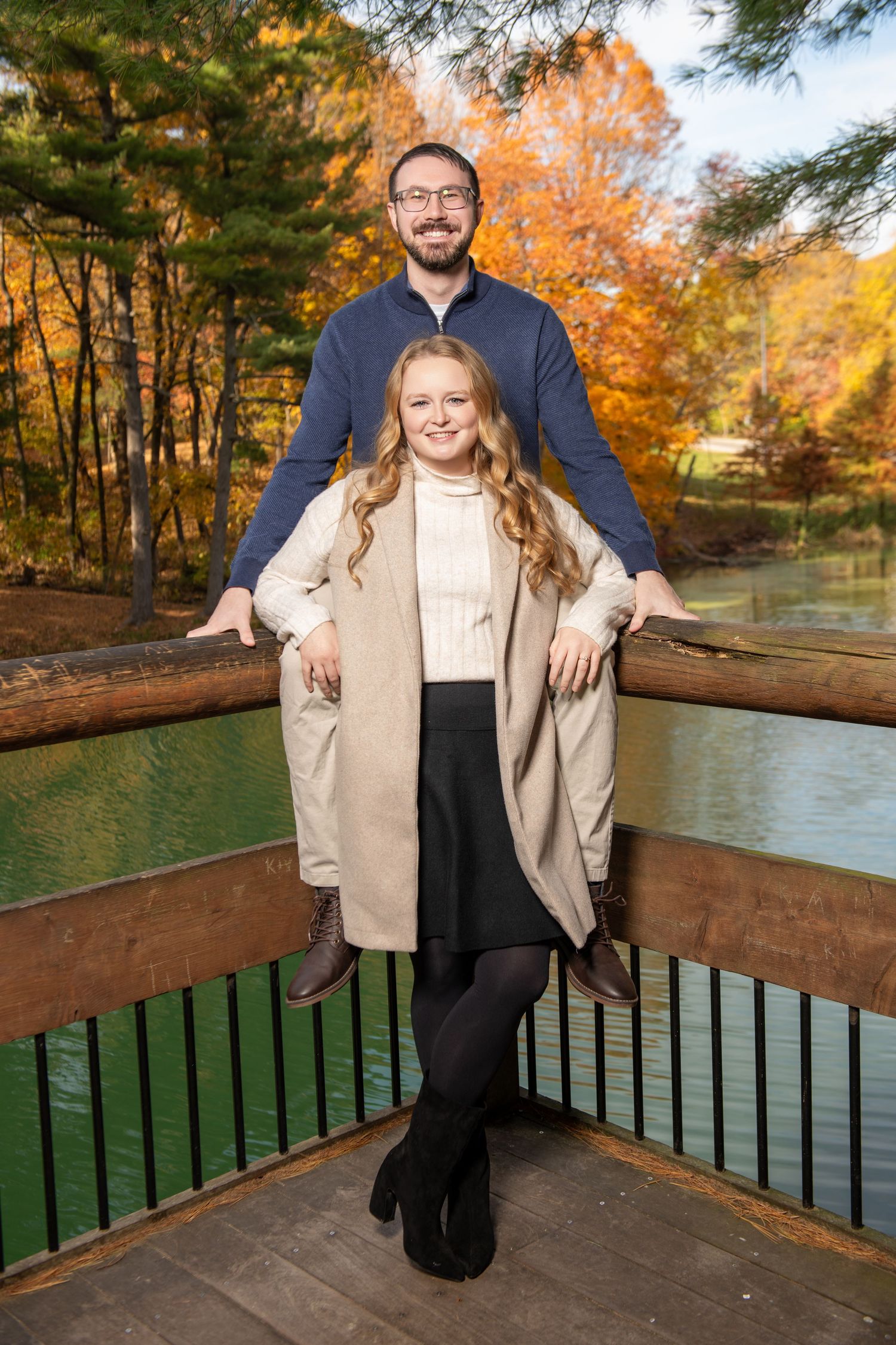 Couple posing on a wooden bridge surrounded by vibrant autumn foliage and a serene green pond.