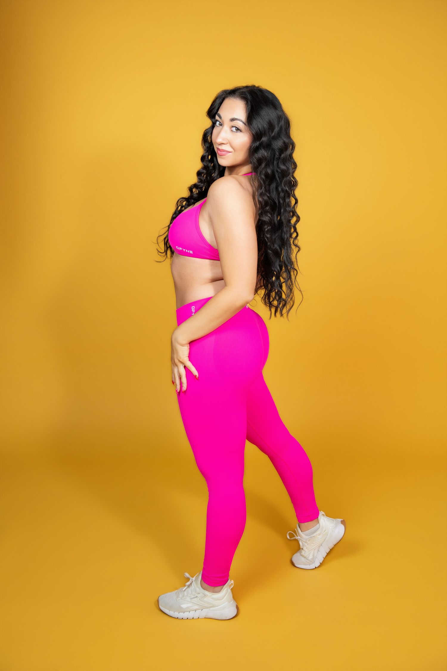 Fit woman in pink sports bra and leggings smiling against bright yellow background.