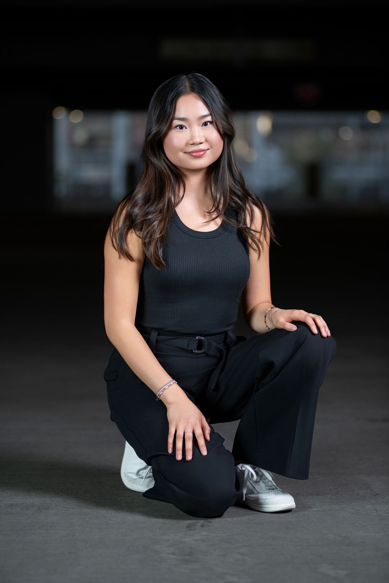 Young Asian woman in all-black outfit crouching and smiling in dimly lit urban setting.
