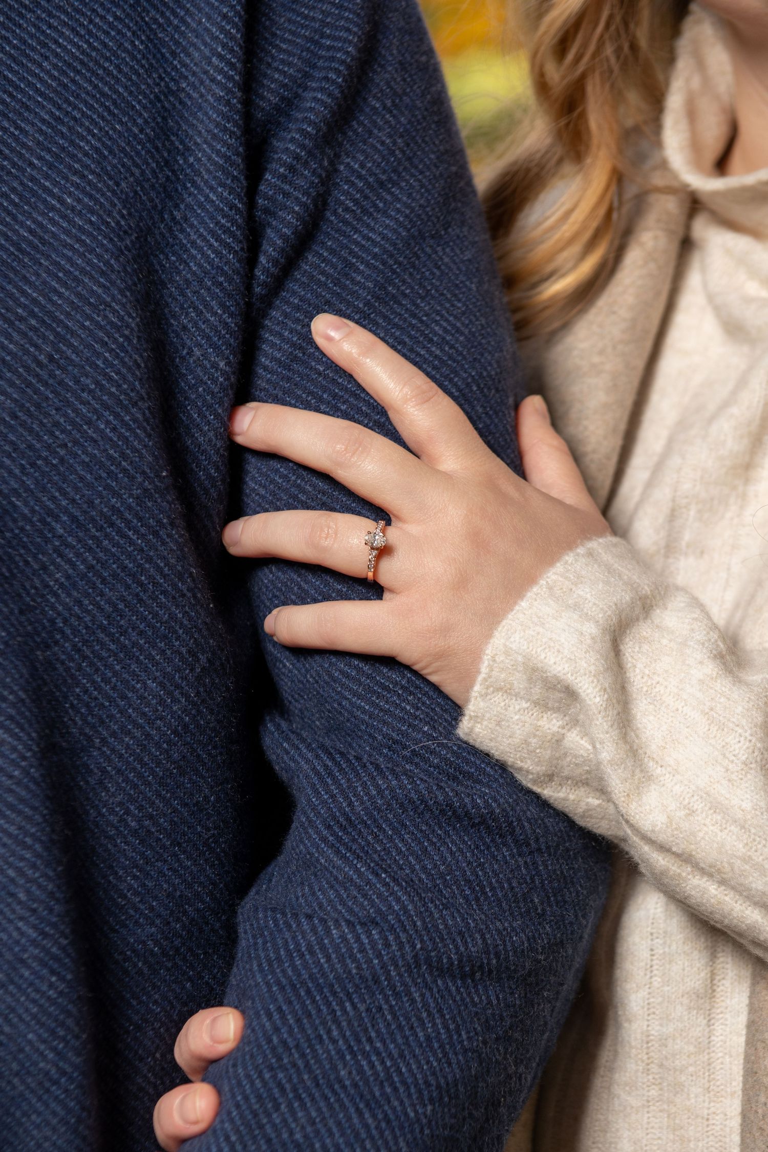 Close-up of woman's hand with diamond engagement ring resting on man's navy blue jacket sleeve.