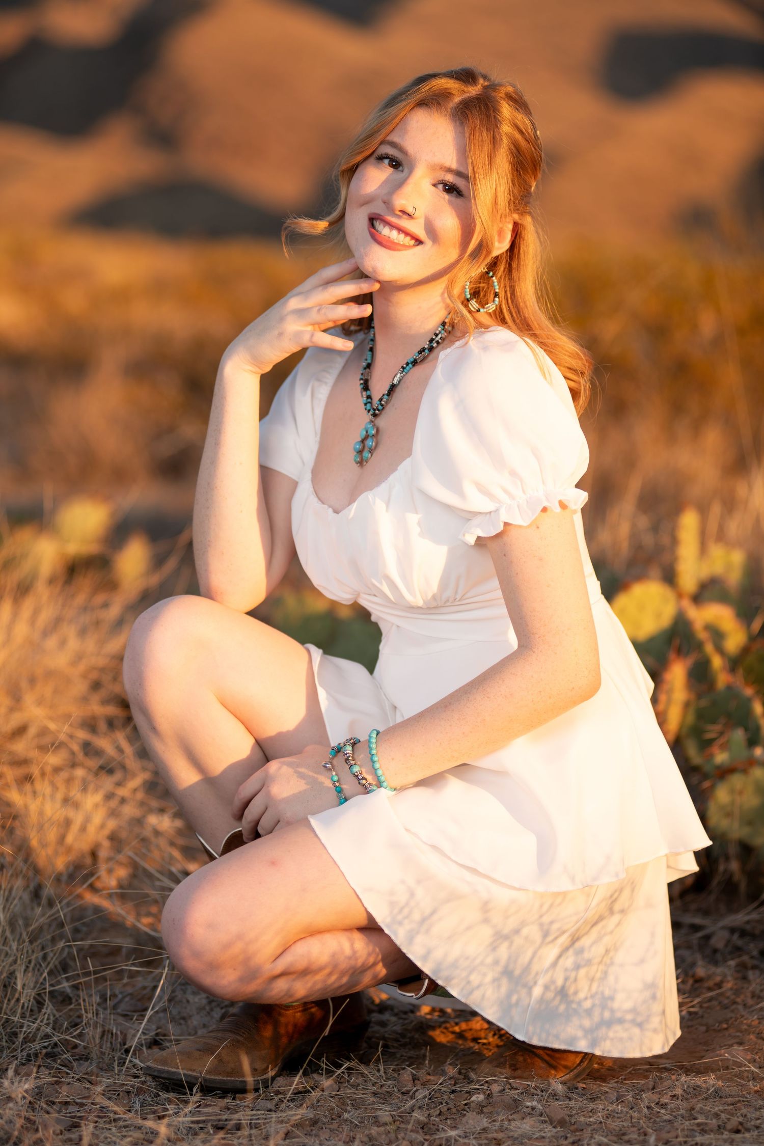 Smiling redhead woman in white dress crouching in desert landscape at golden hour.